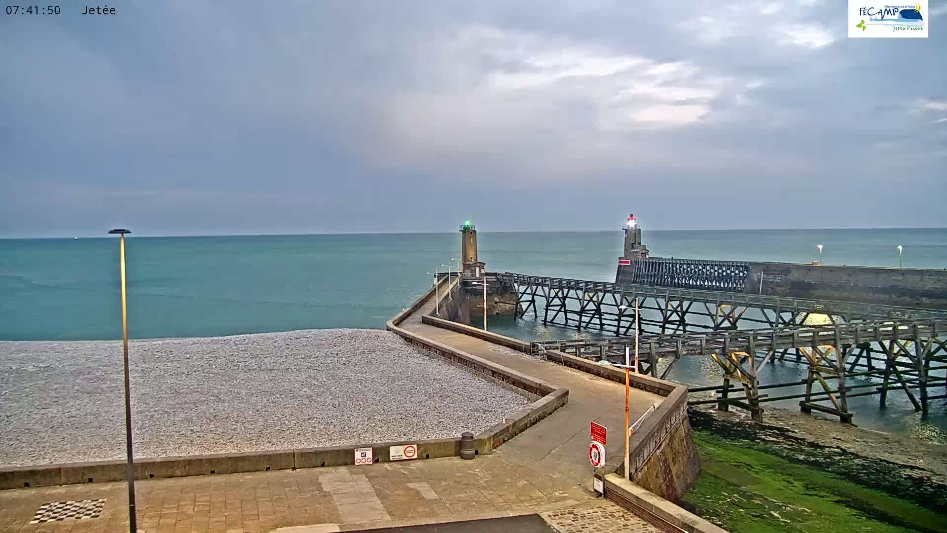 A long pier with two lighthouses extends into a calm, teal ocean under an overcast sky, with a pebble beach visible to the left.