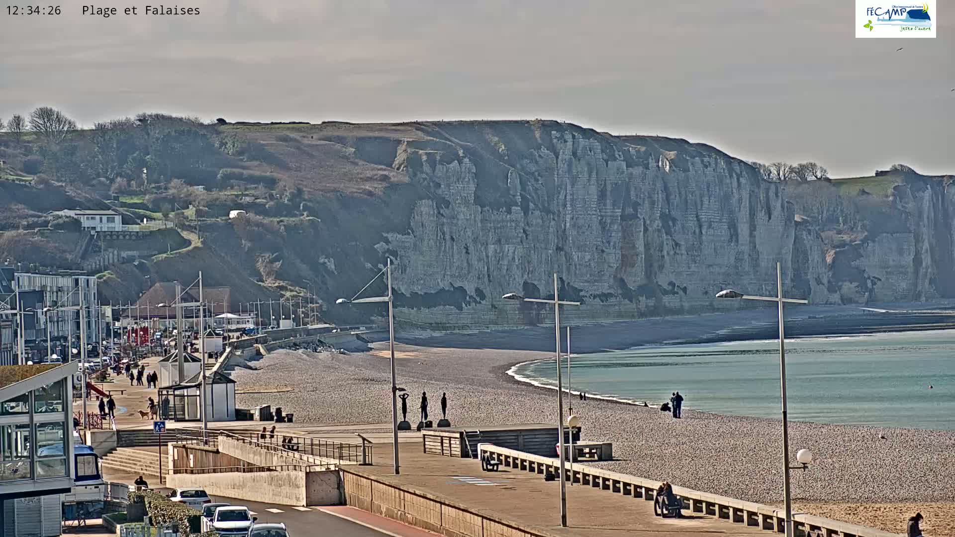 A pebble beach and boardwalk with a few people, overlooking a large, light-grey cliff face under a mostly cloudy sky.