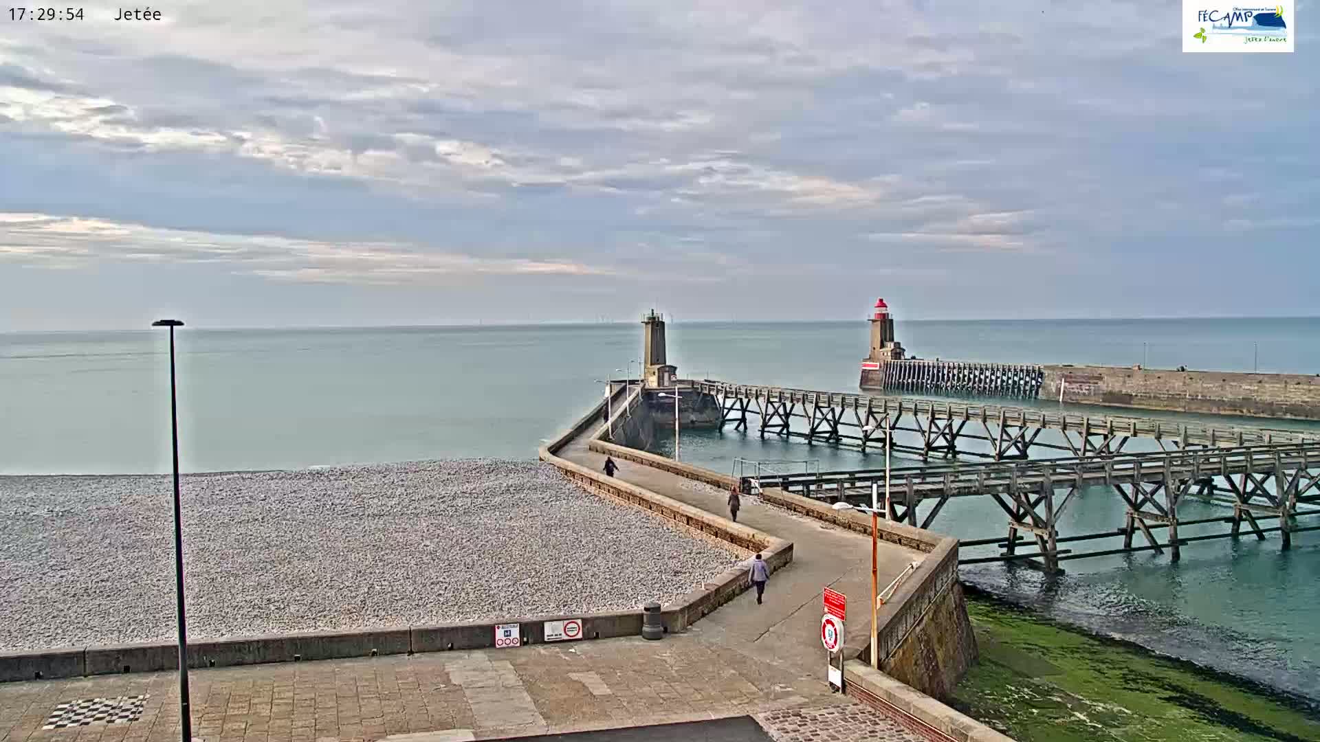 A pebble beach stretches toward two lighthouses and a long wooden pier under a partly cloudy sky.