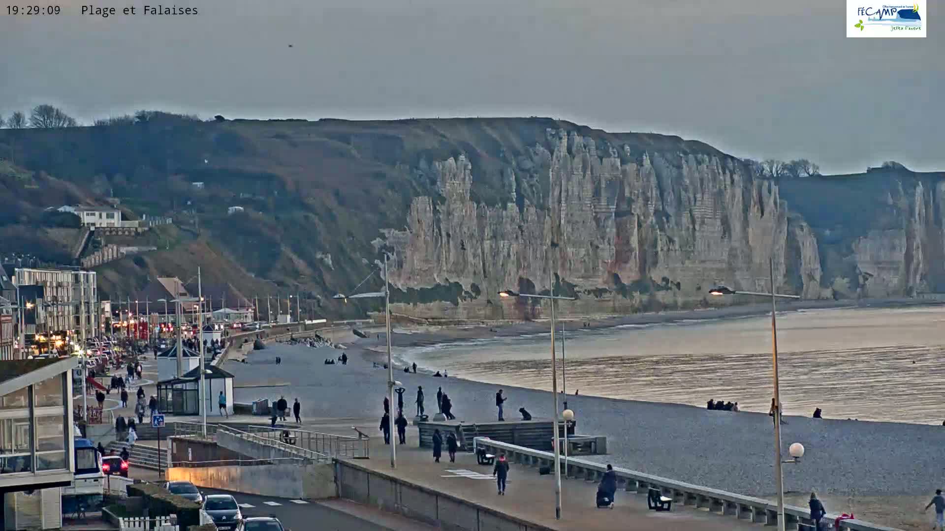 A beach with a pebble shoreline and people walking along a promenade, overlooks a large cliff face under an overcast sky.