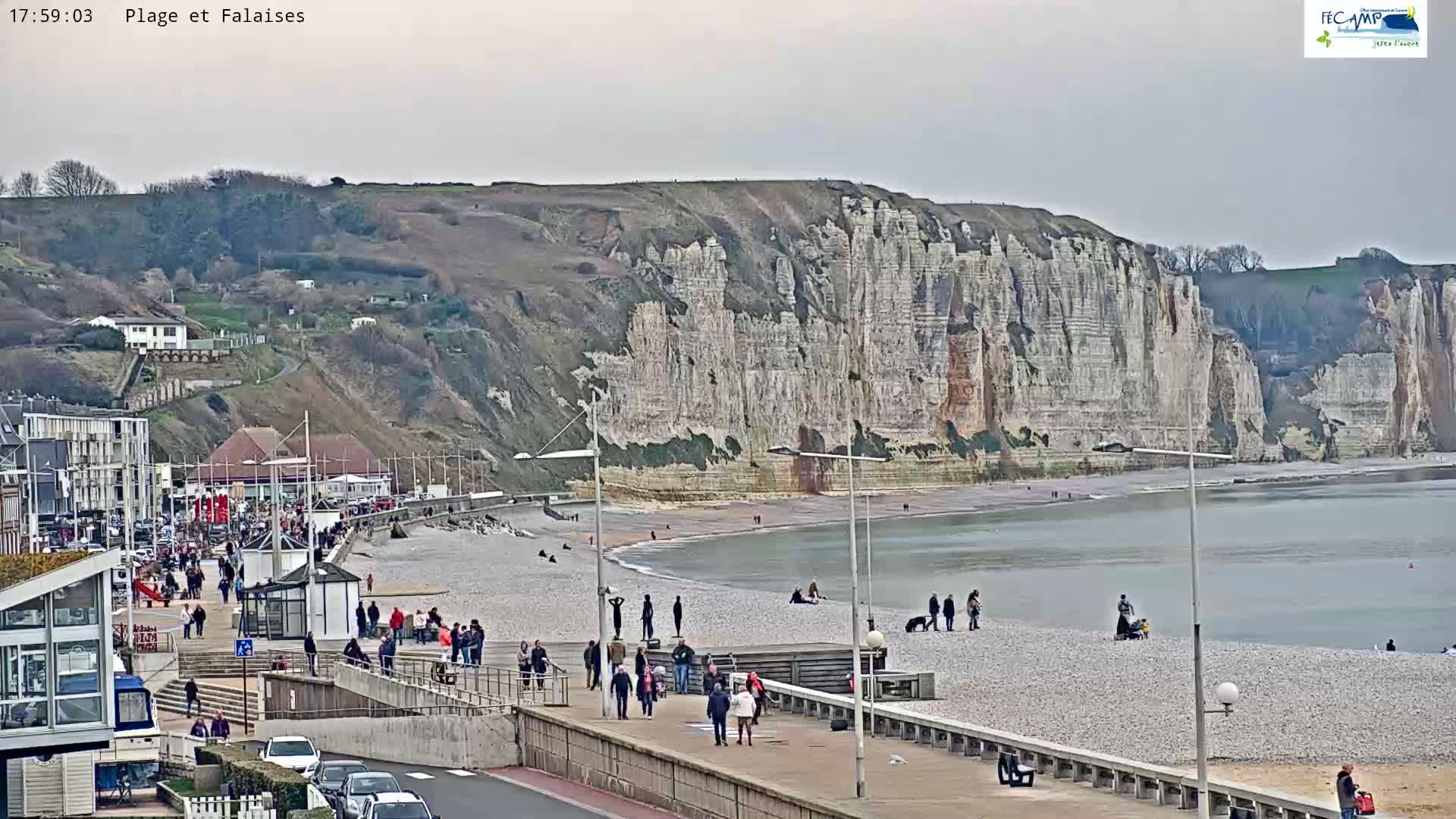 A pebble beach with people walking along a boardwalk, overlooking a large expanse of white cliffs under an overcast sky.