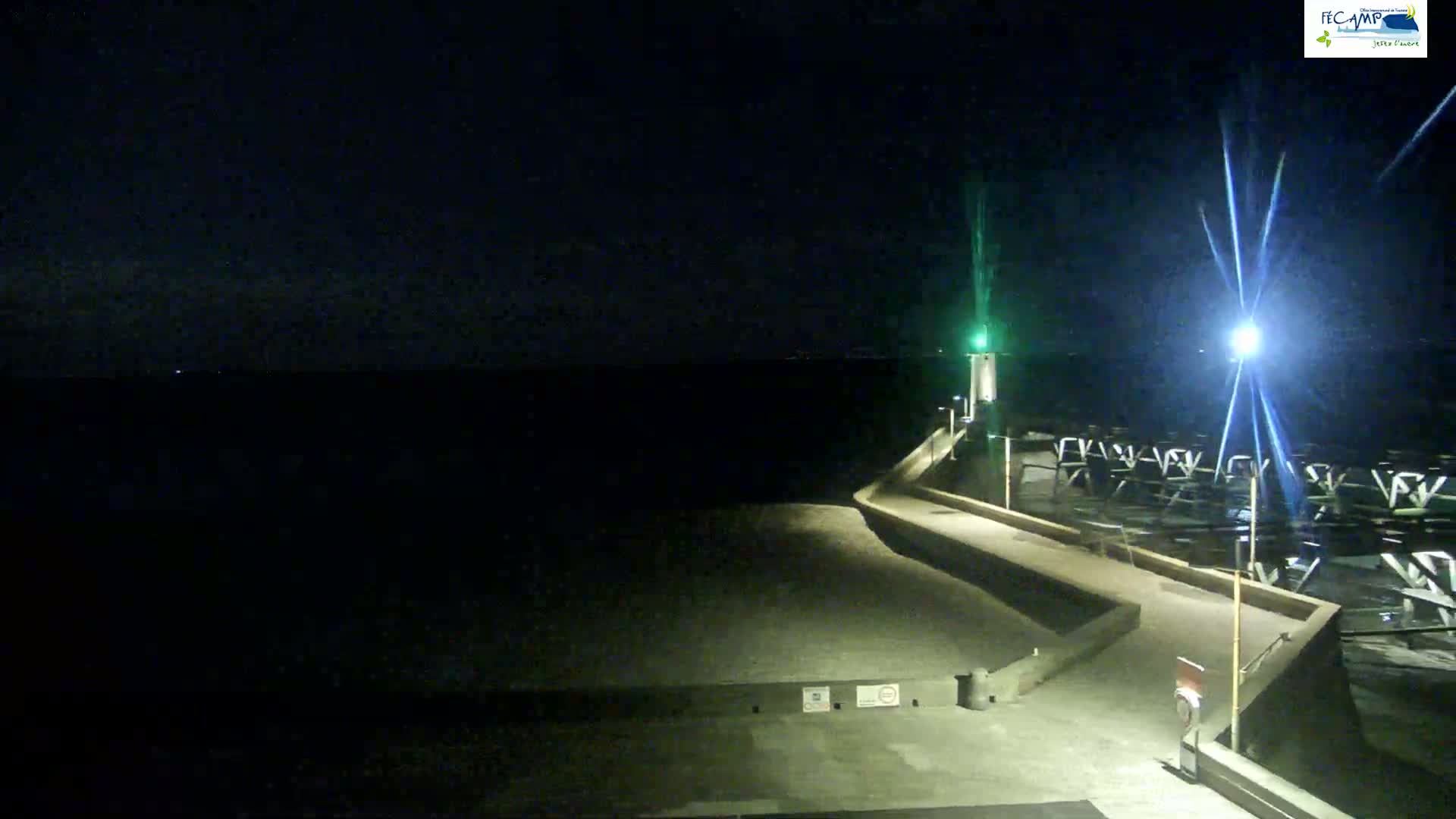 A nighttime view of a dark, calm body of water next to a lit walkway and pier, with a small lighthouse visible in the distance.