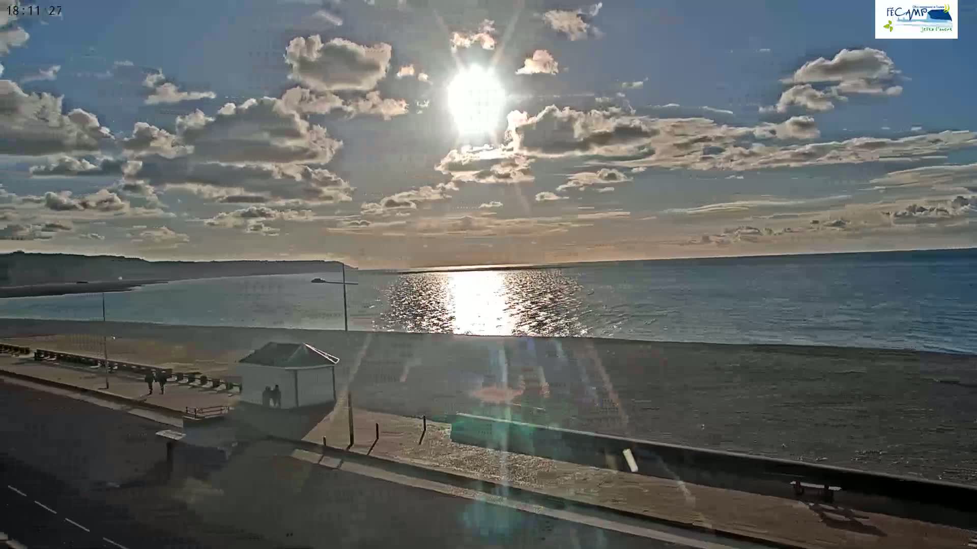 A partly cloudy sunset shines over a calm ocean beach with a small building and several people visible along the shoreline.