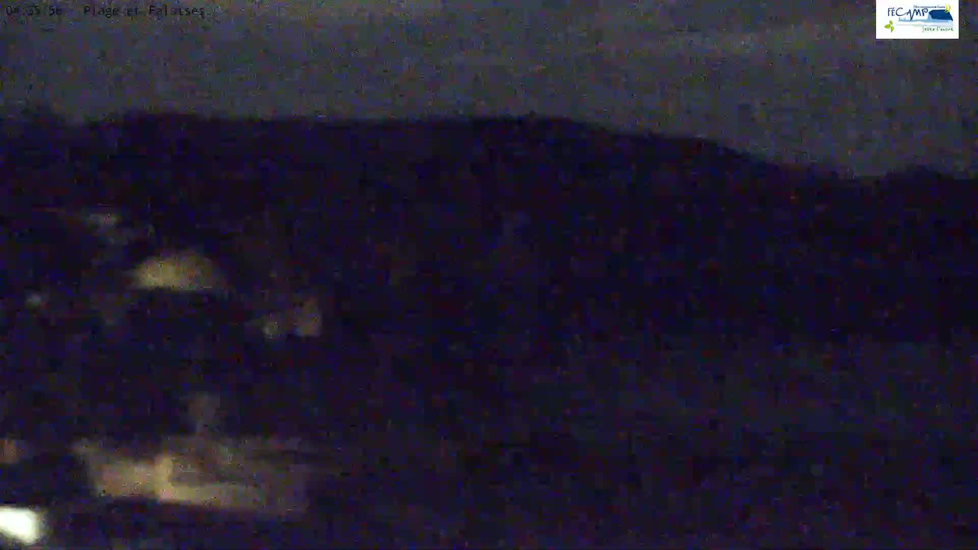 A dark, nighttime scene shows a body of water with faint, blurry lights near the bottom, and a dark shoreline in the background under a mostly cloudy sky.