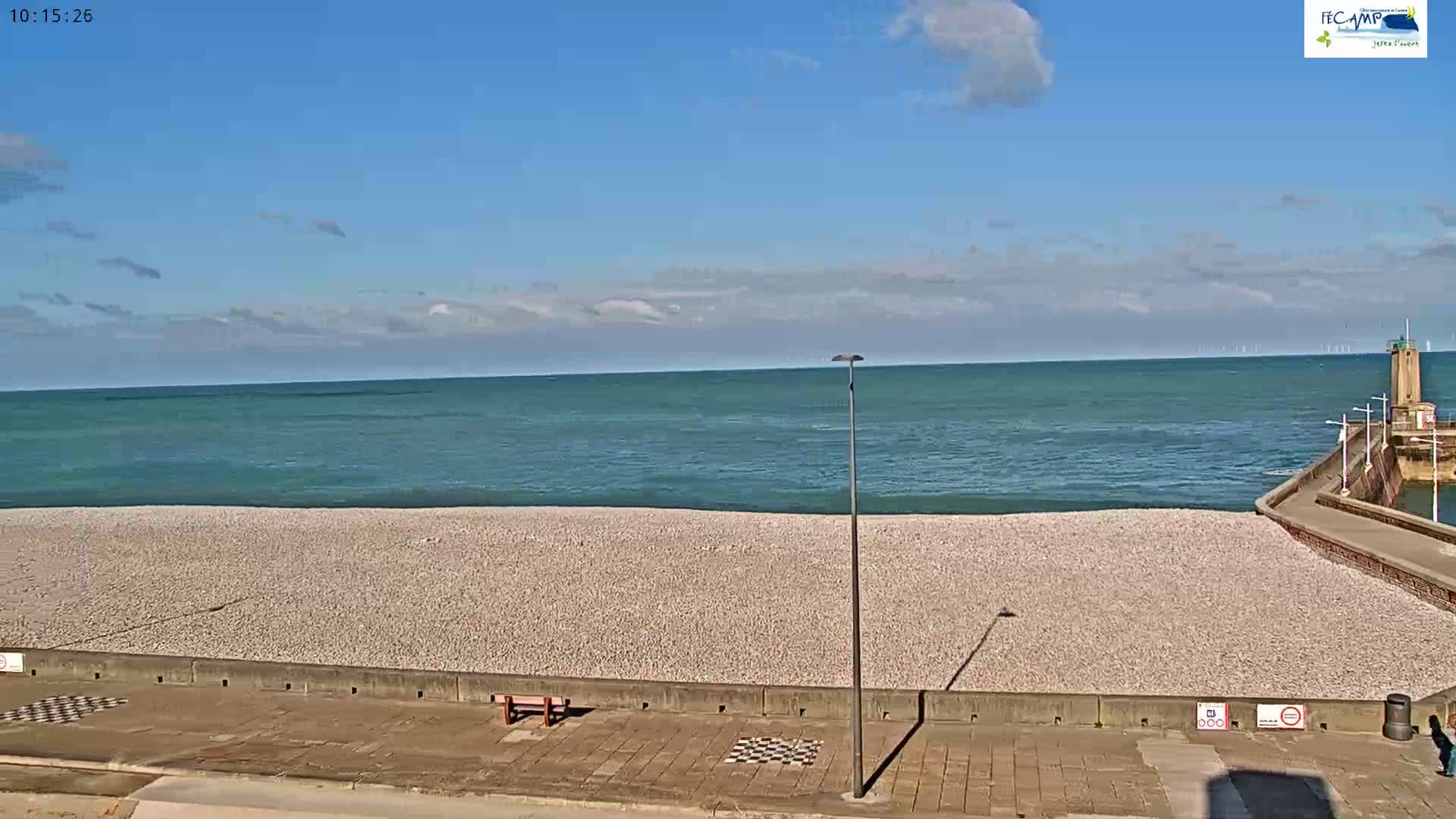 A pebble beach stretches along a calm, teal ocean under a mostly clear, blue sky.