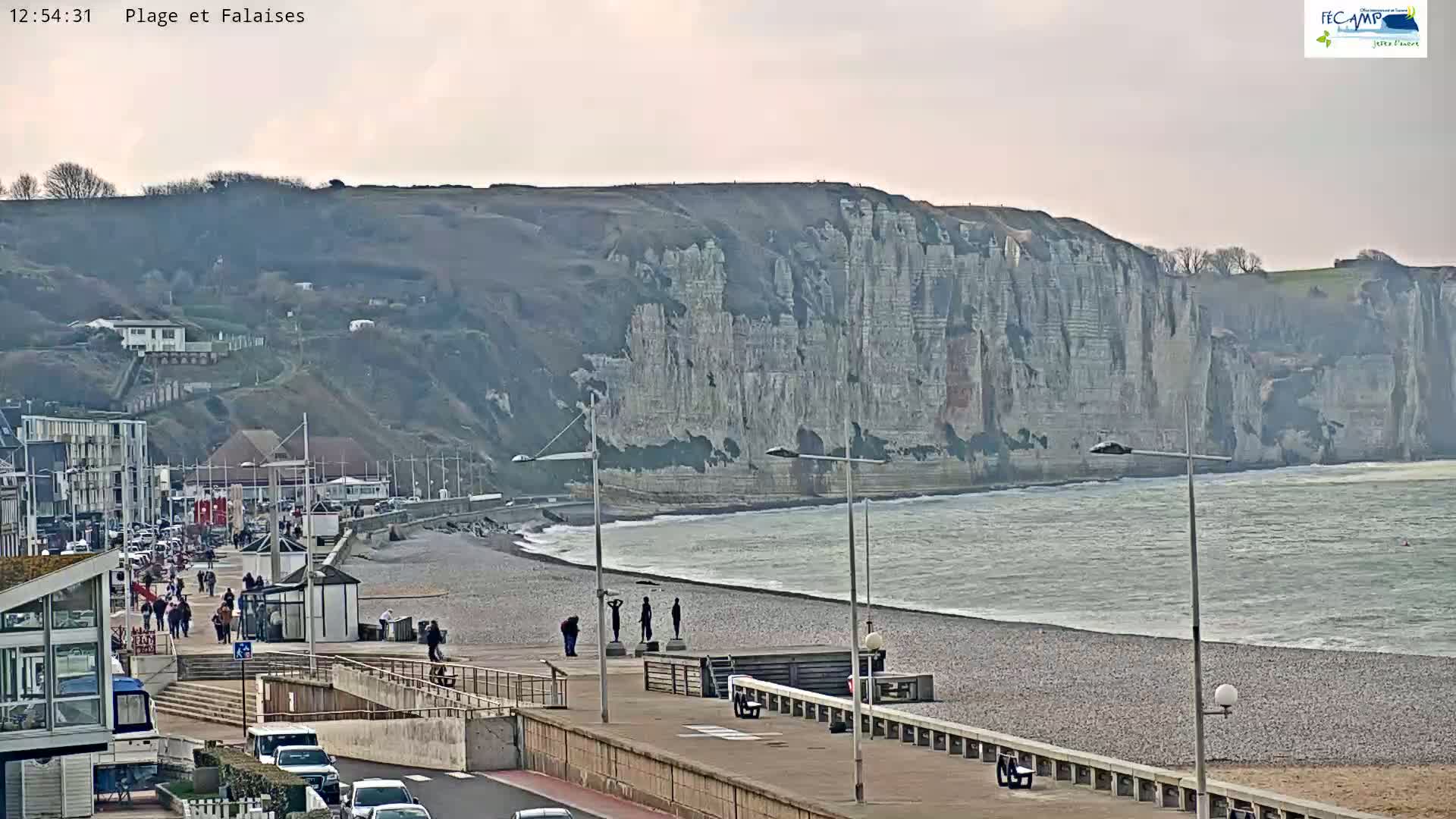 A pebble beach with people walking along a promenade, overlooking a calm ocean and towering white cliffs under an overcast sky.