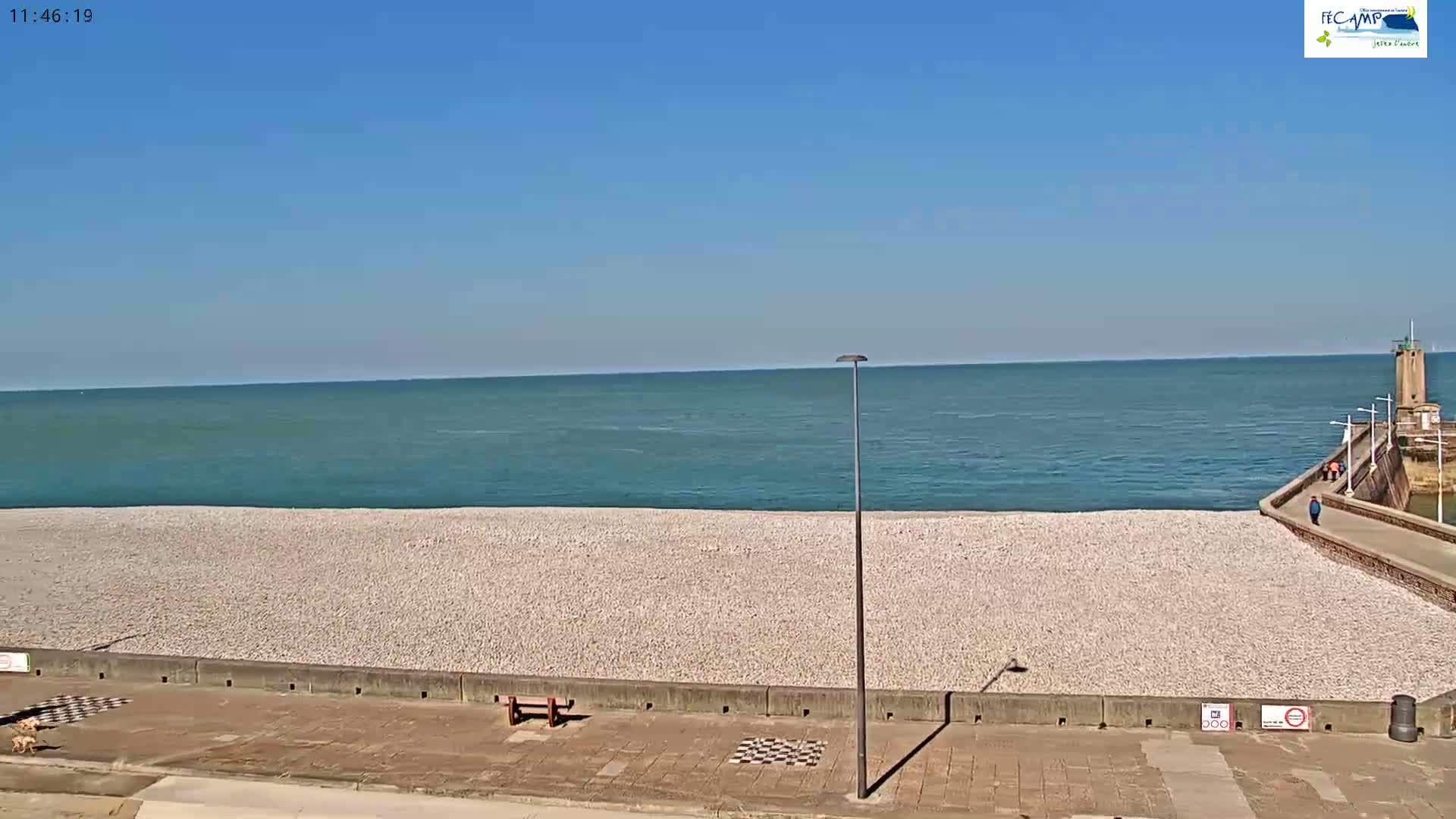 A mostly sunny day shows a pebble beach with a calm teal ocean, a walkway, and a small tower in the distance.
