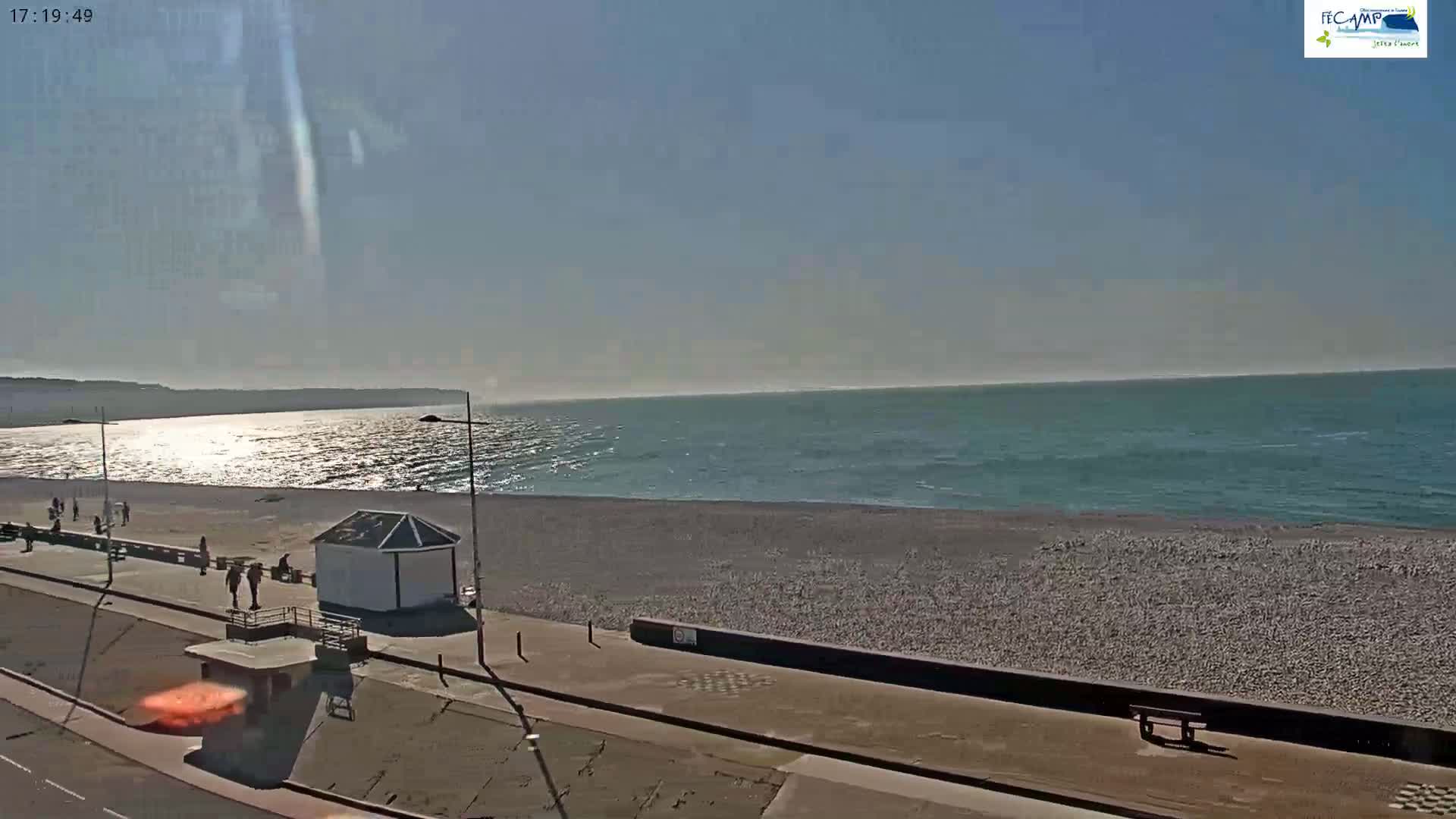 A mostly sunny day shows a pebble beach and calm ocean with a few people walking along the shoreline promenade, near a small building.