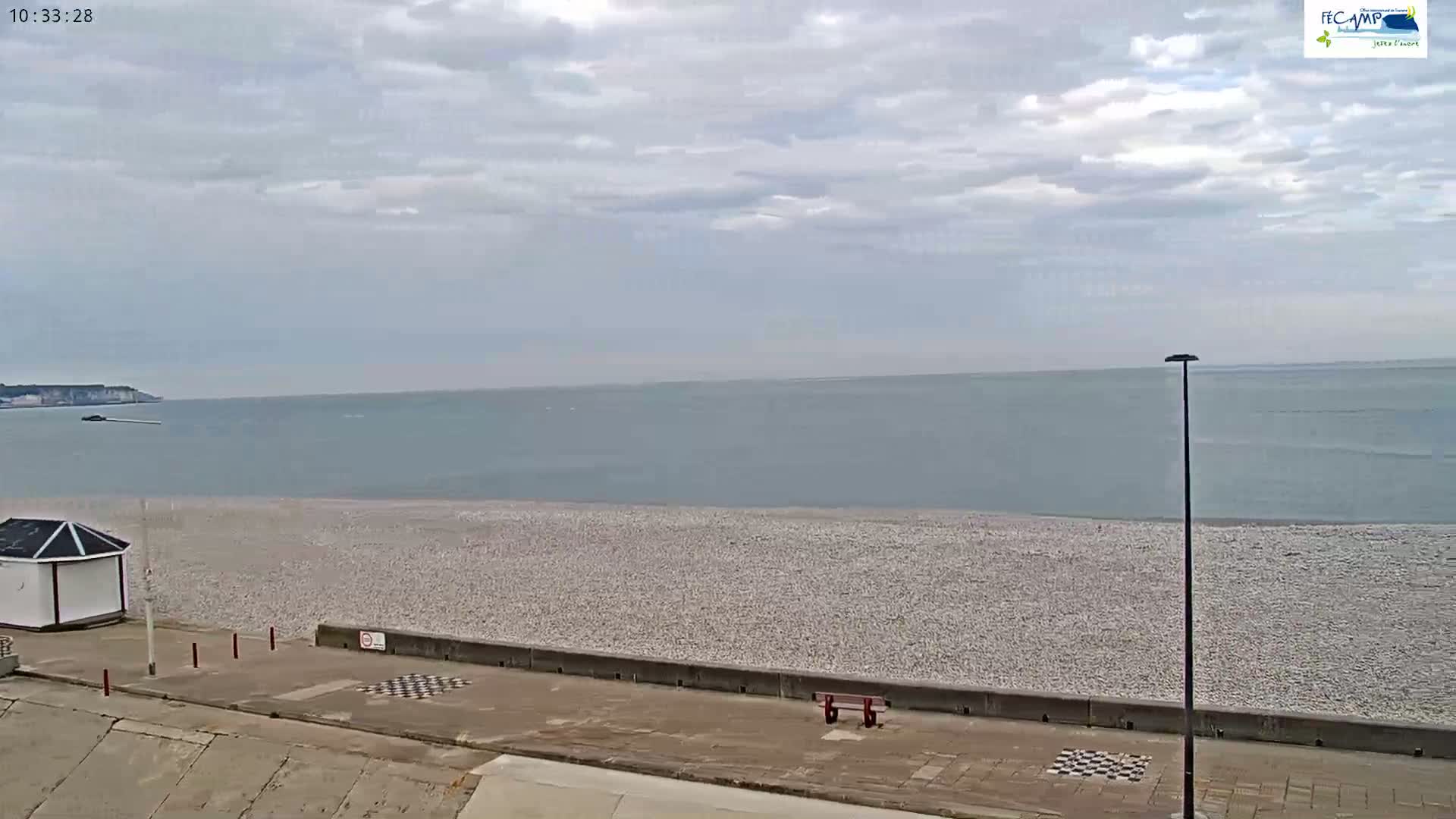 A pebble beach stretches to a calm sea under a mostly cloudy sky, with a small white building and a red bench visible in the foreground.