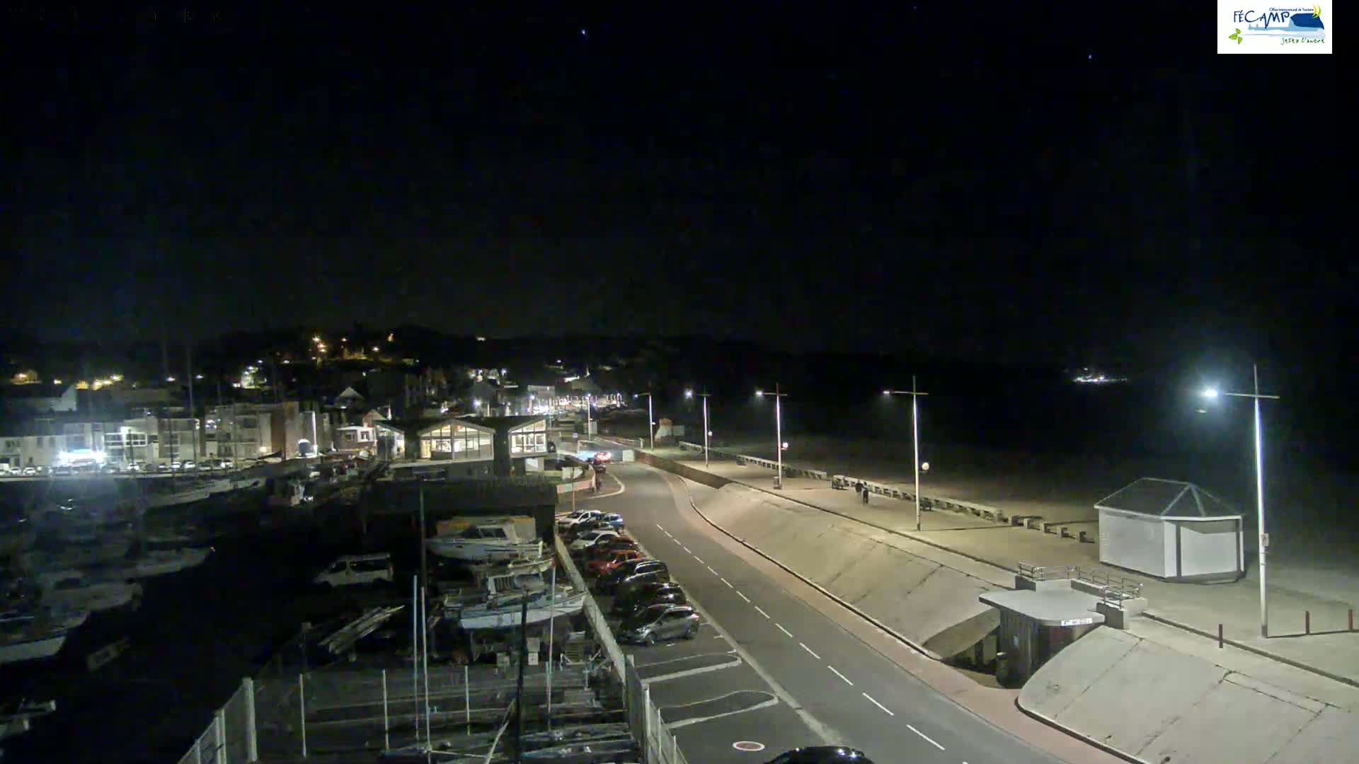 A nighttime, high-angle view shows a coastal town with a harbor full of boats, a road leading to a beach with people walking along the promenade under bright streetlights.