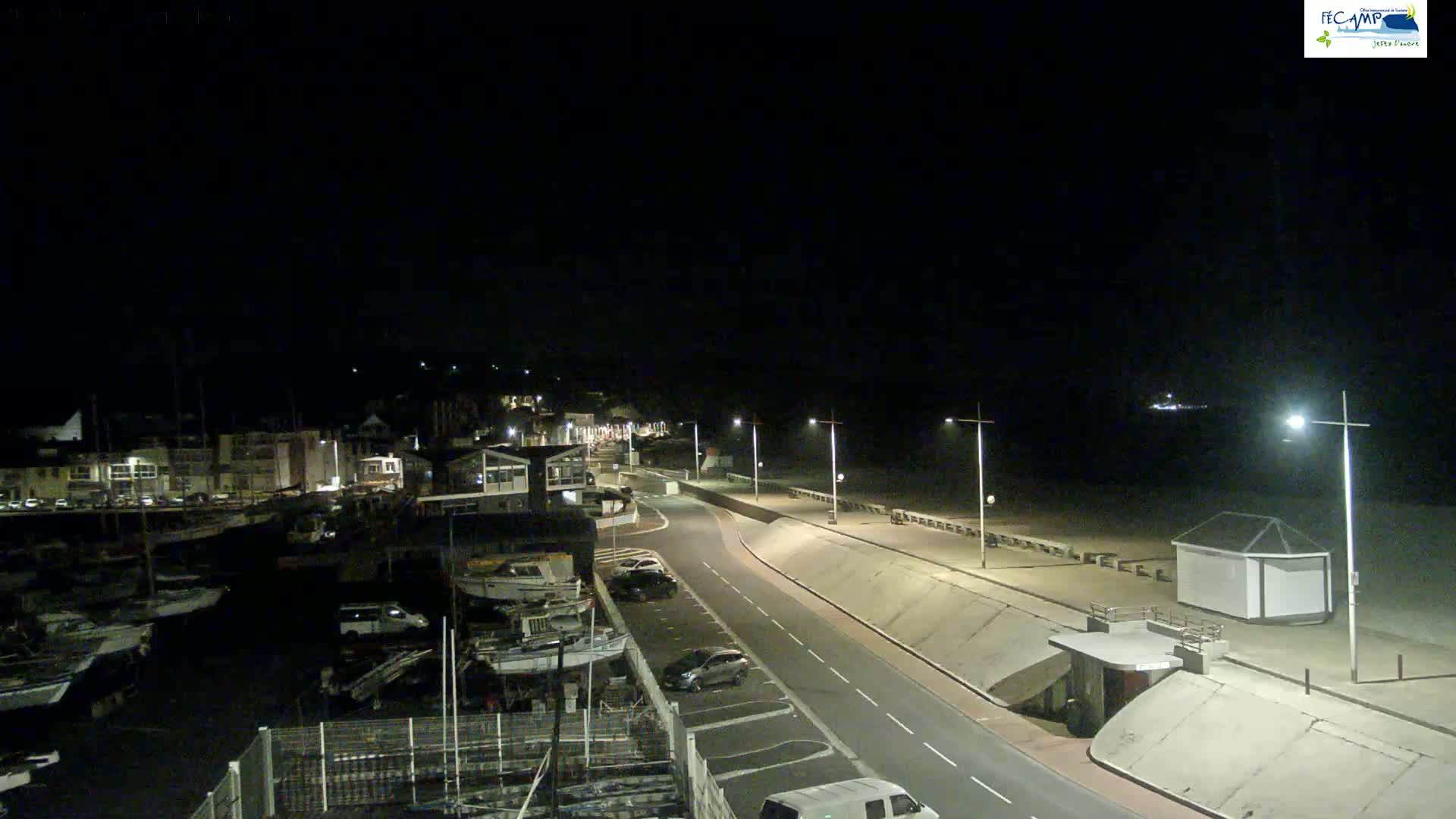 A nighttime, coastal scene shows a road curving along a seawall toward a beach, with a harbor full of boats on the left and buildings lining the road.