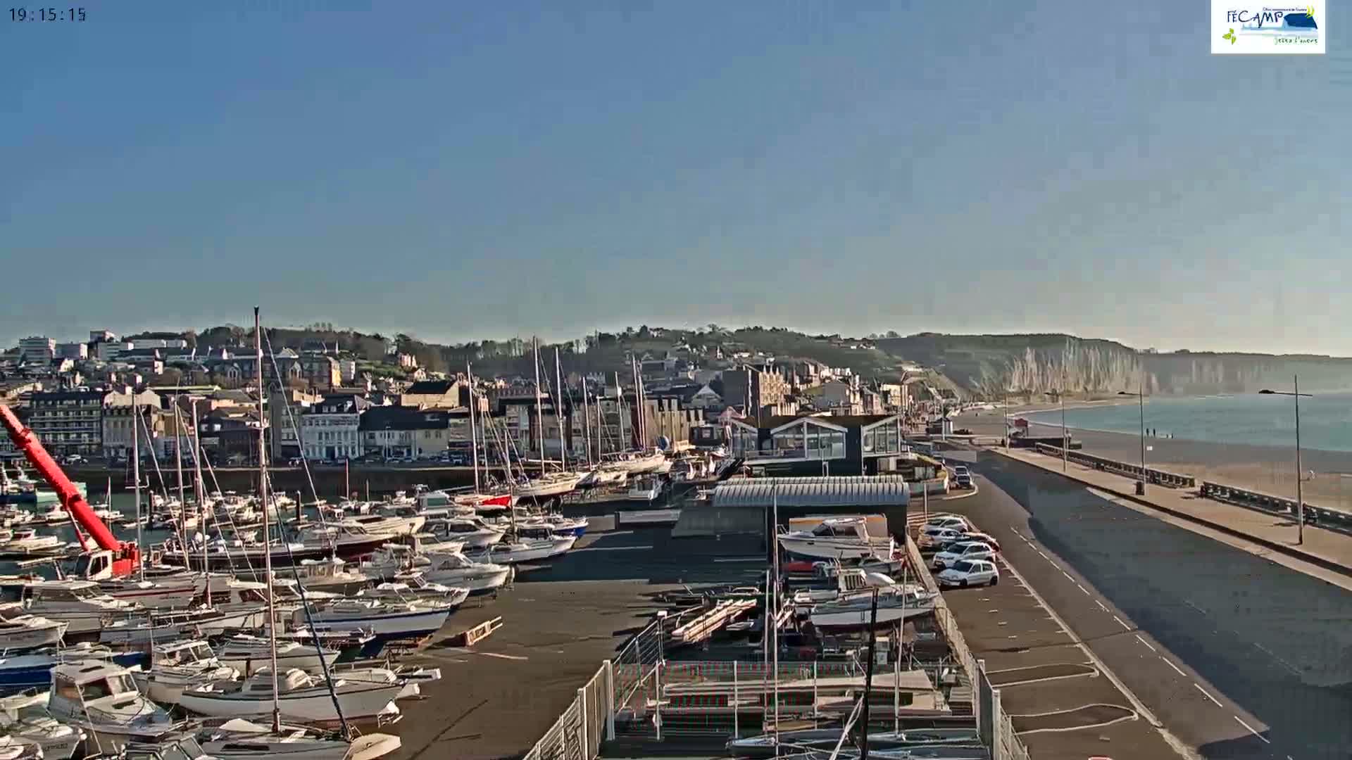 A harbor filled with numerous sailboats and motorboats is situated next to a town, with a beach and high white cliffs visible in the background under a clear, sunny sky.