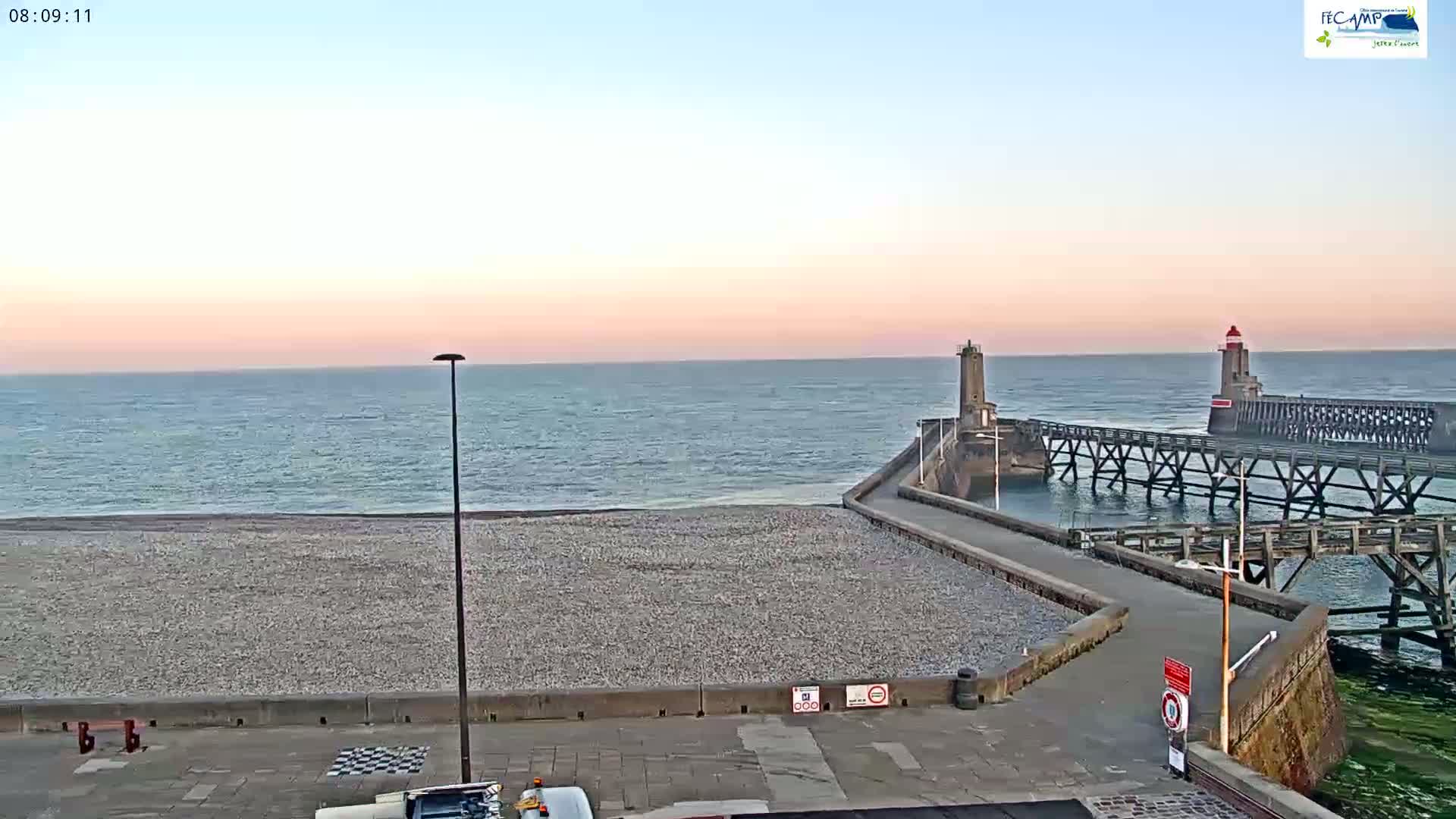 A pebble beach and a long pier with two lighthouses extend into a calm, twilight ocean under a pale pink sky.