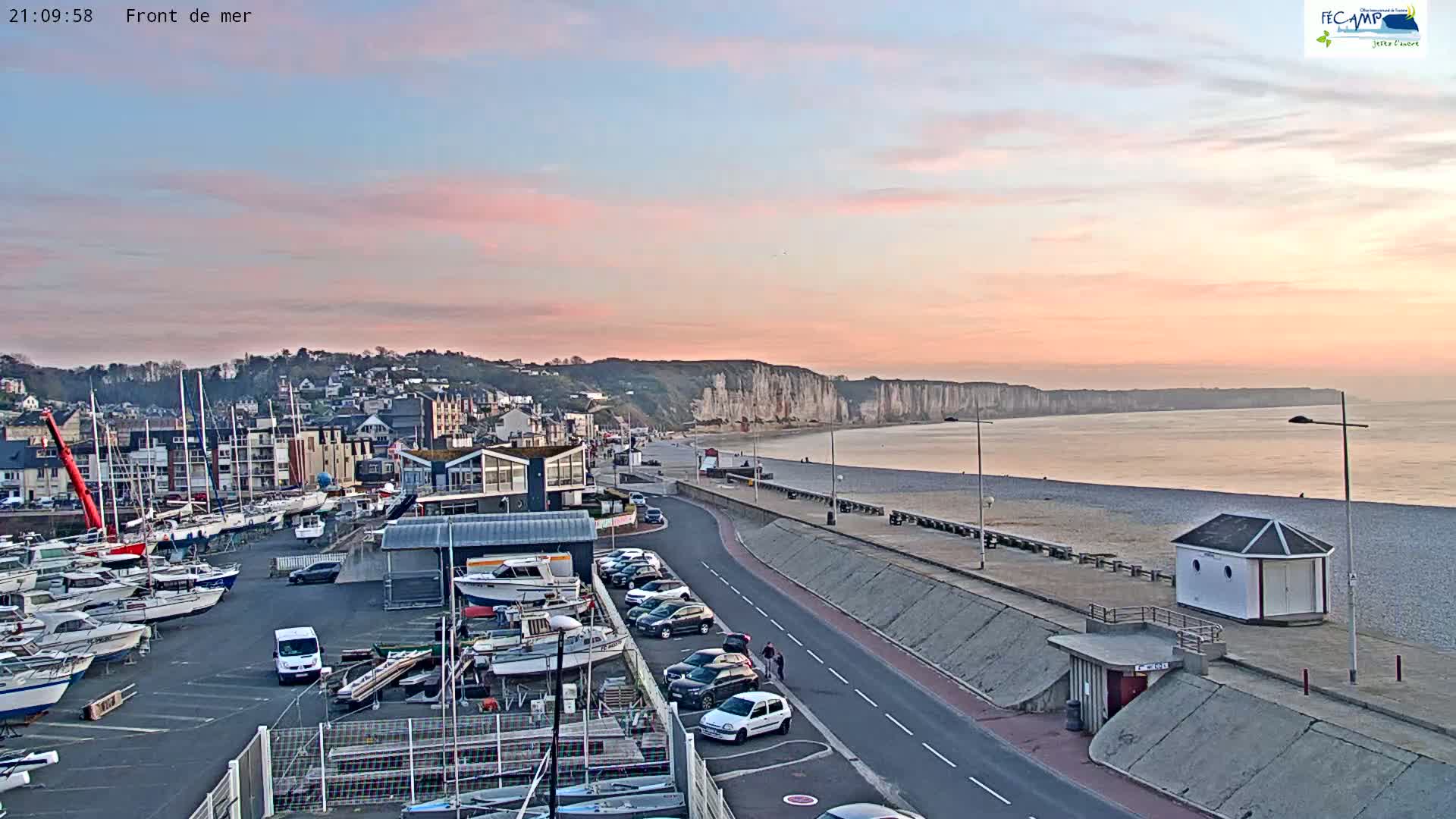 A coastal town at sunset, showing a marina filled with boats, a road along a pebble beach, and high white cliffs in the distance.