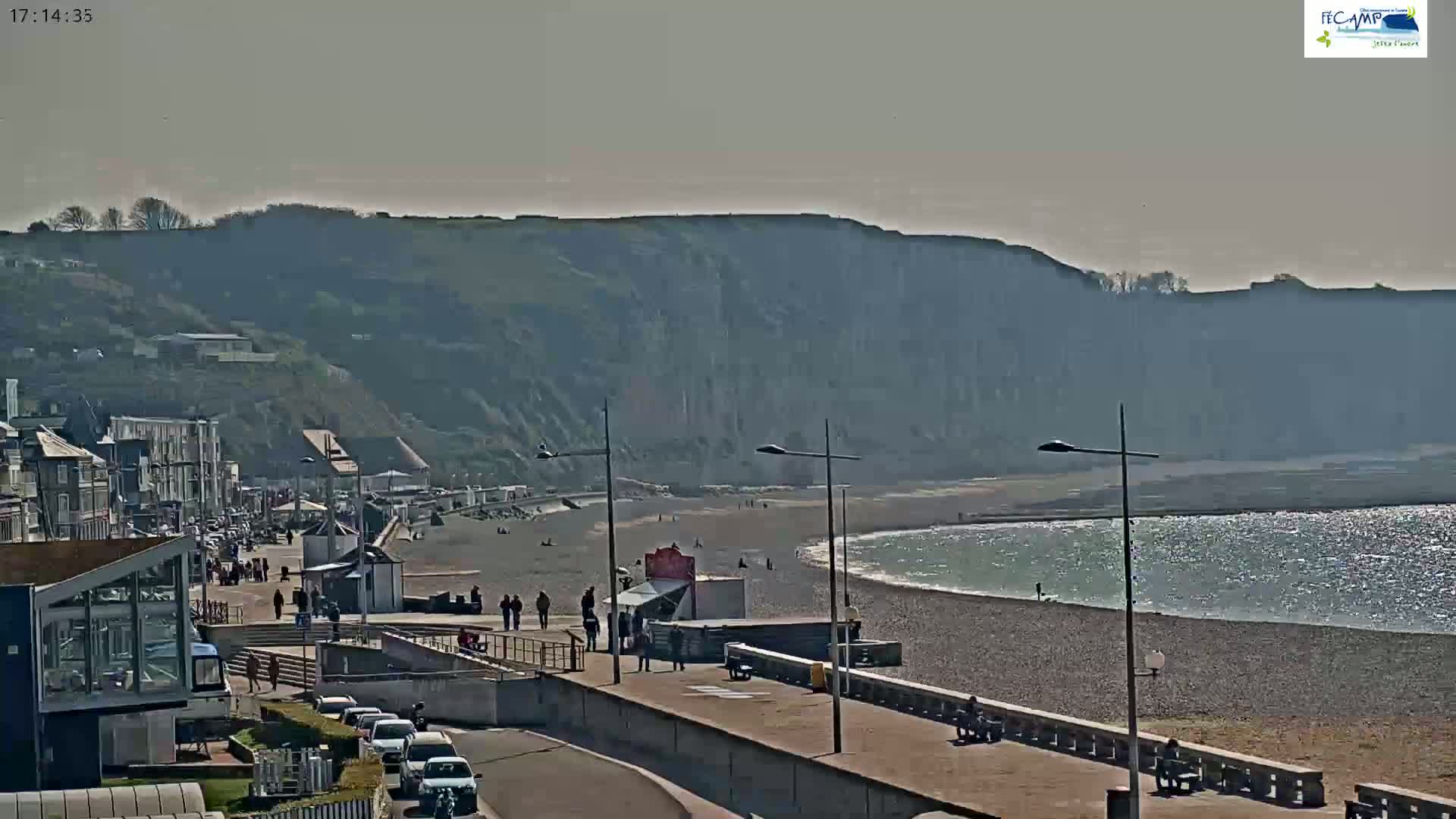 A mostly sunny day shows a pebble beach and calm ocean next to a town with buildings and a promenade, all situated below a large, light-grey cliff.