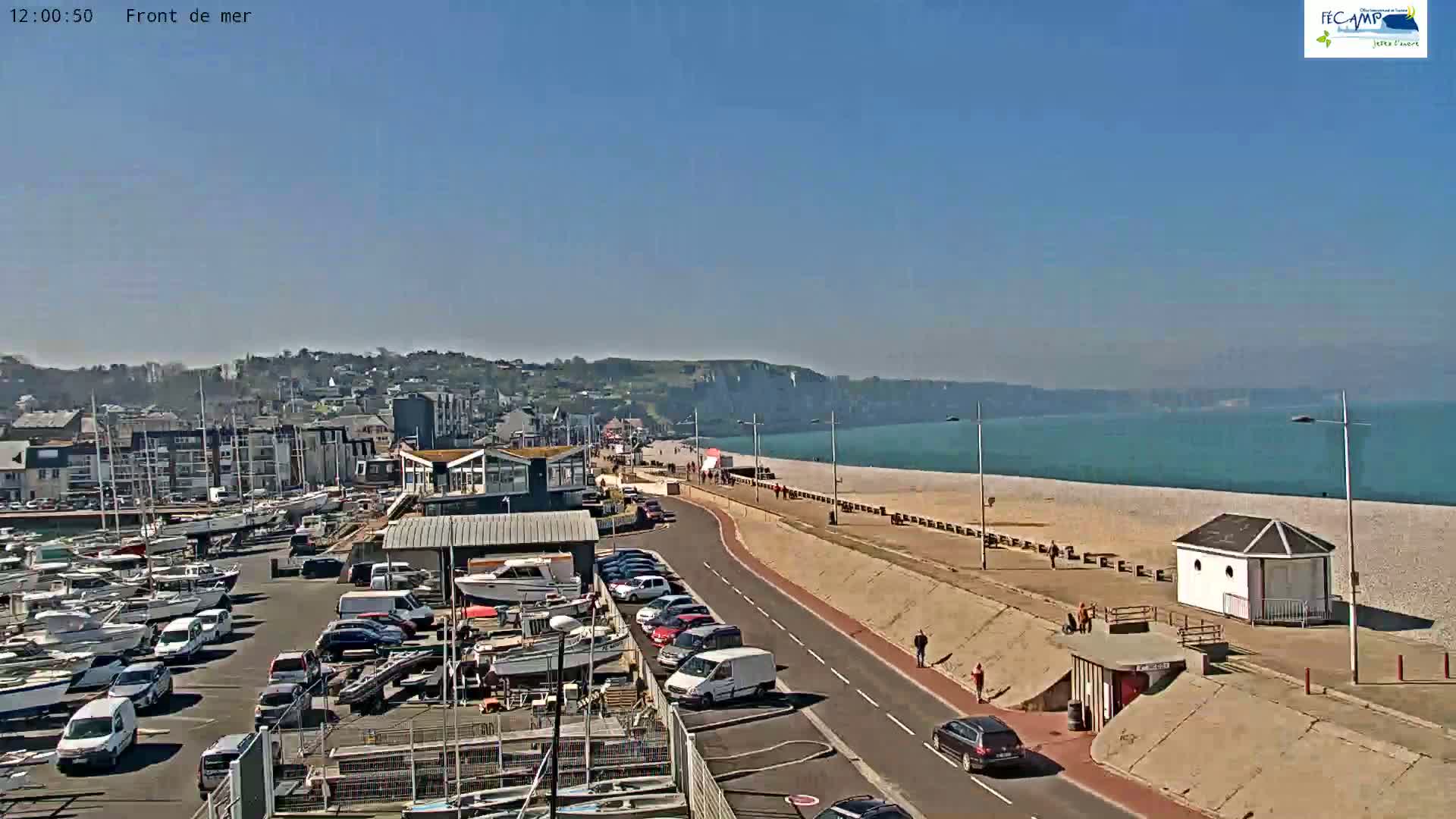 A sunny day overlooking a harbor filled with boats, a seaside promenade with pedestrians, and a pebble beach bordering a calm ocean.