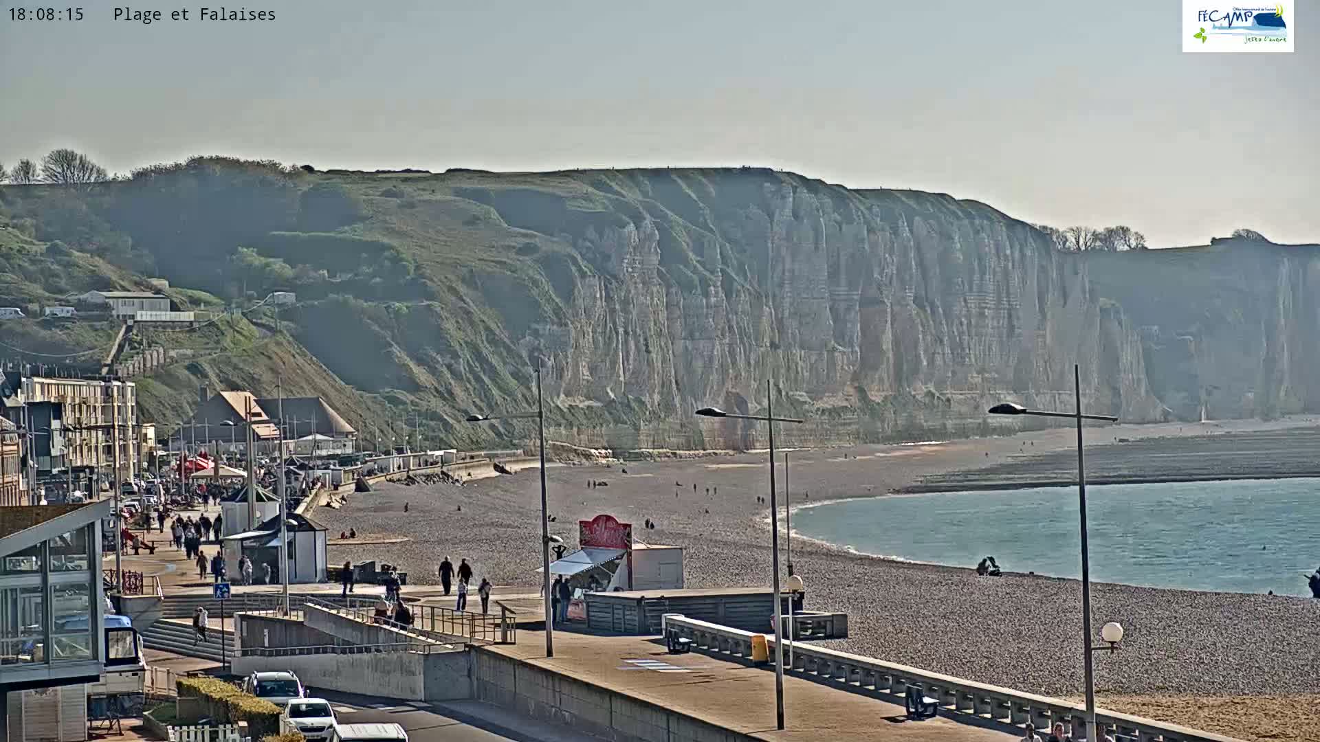 A pebble beach with people walking along a promenade, overlooking a calm ocean and towering white cliffs under a mostly clear sky.