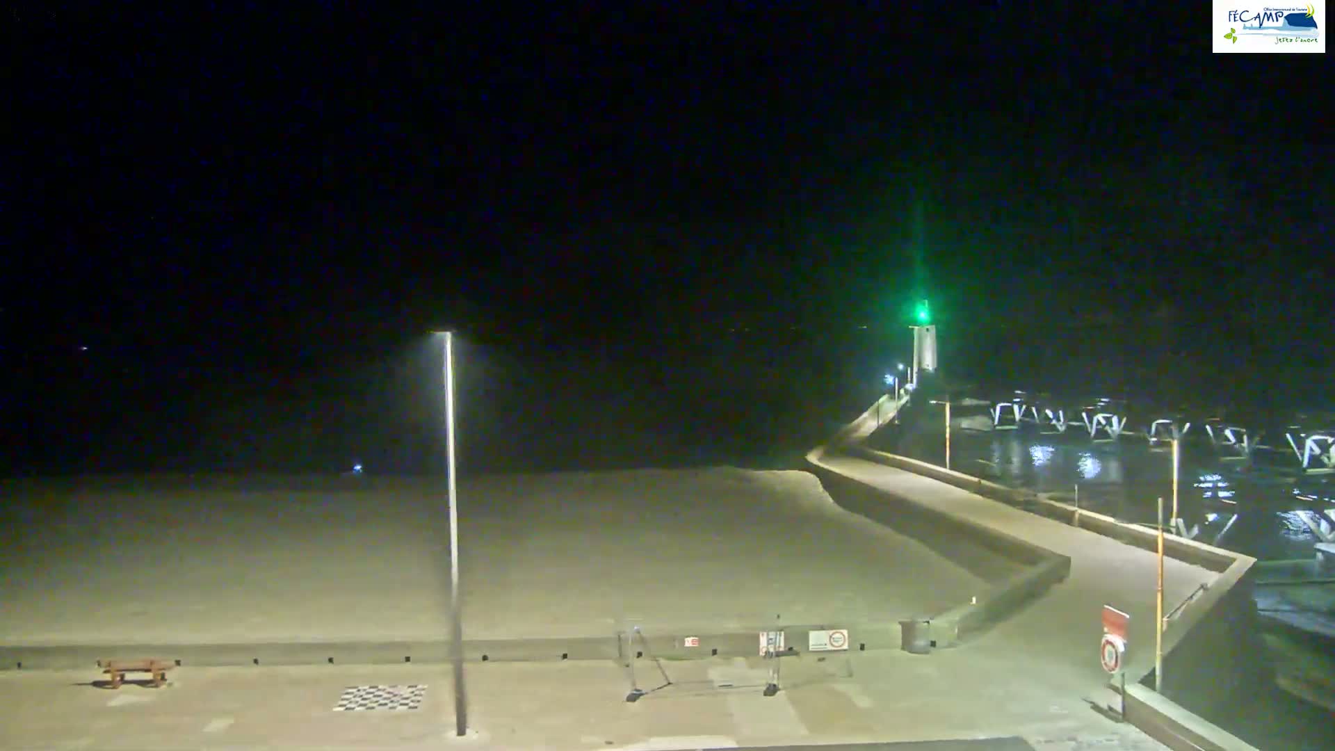 A nighttime view shows a beach, a walkway leading to a harbor with a lit lighthouse, under a clear, dark sky.