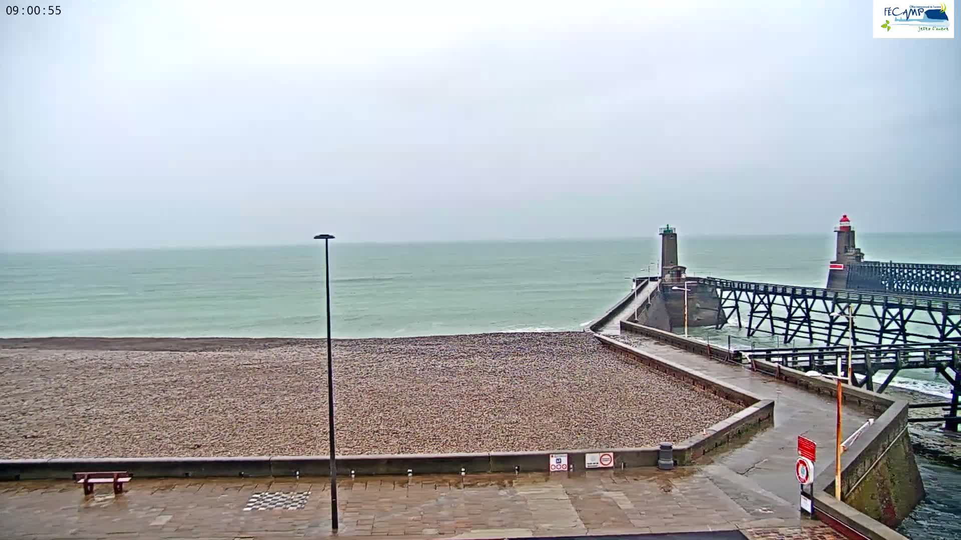 On an overcast day with wet ground, a pebble beach and promenade front a green-grey sea, flanked by a pier with two lighthouses extending into the water.