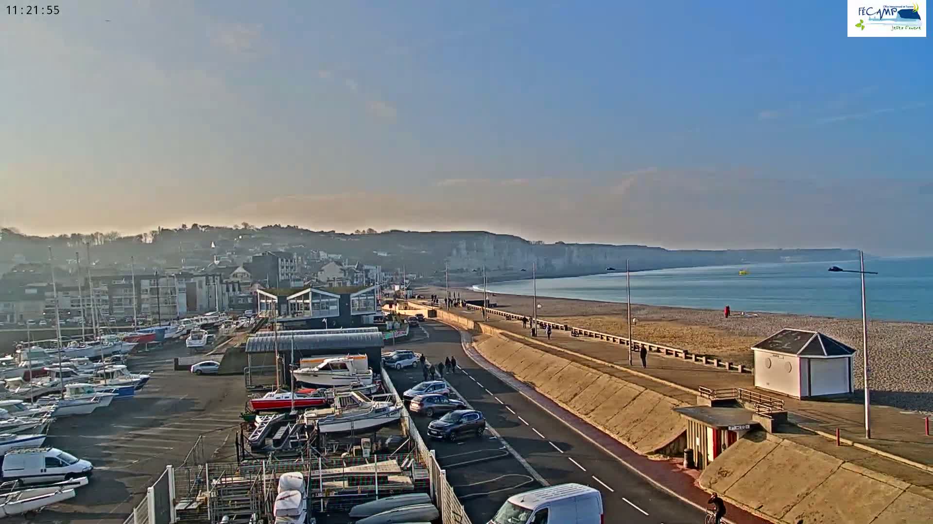 On an overcast day with wet ground, a pebble beach and promenade front a green-grey sea, flanked by a pier with two lighthouses extending into the water.