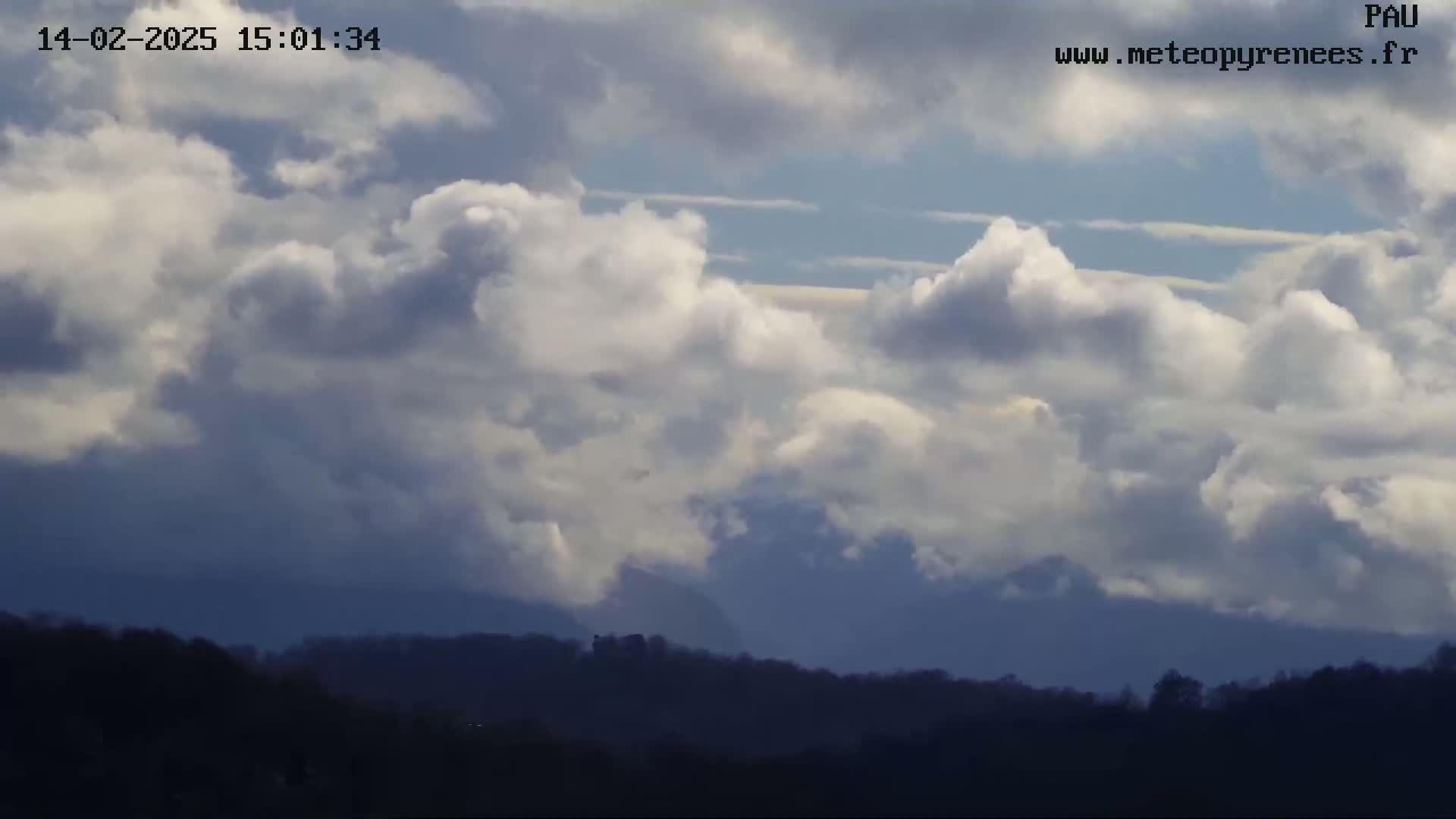 A partly cloudy sky with fluffy white and gray clouds hangs over a dark, silhouetted line of trees and hills.