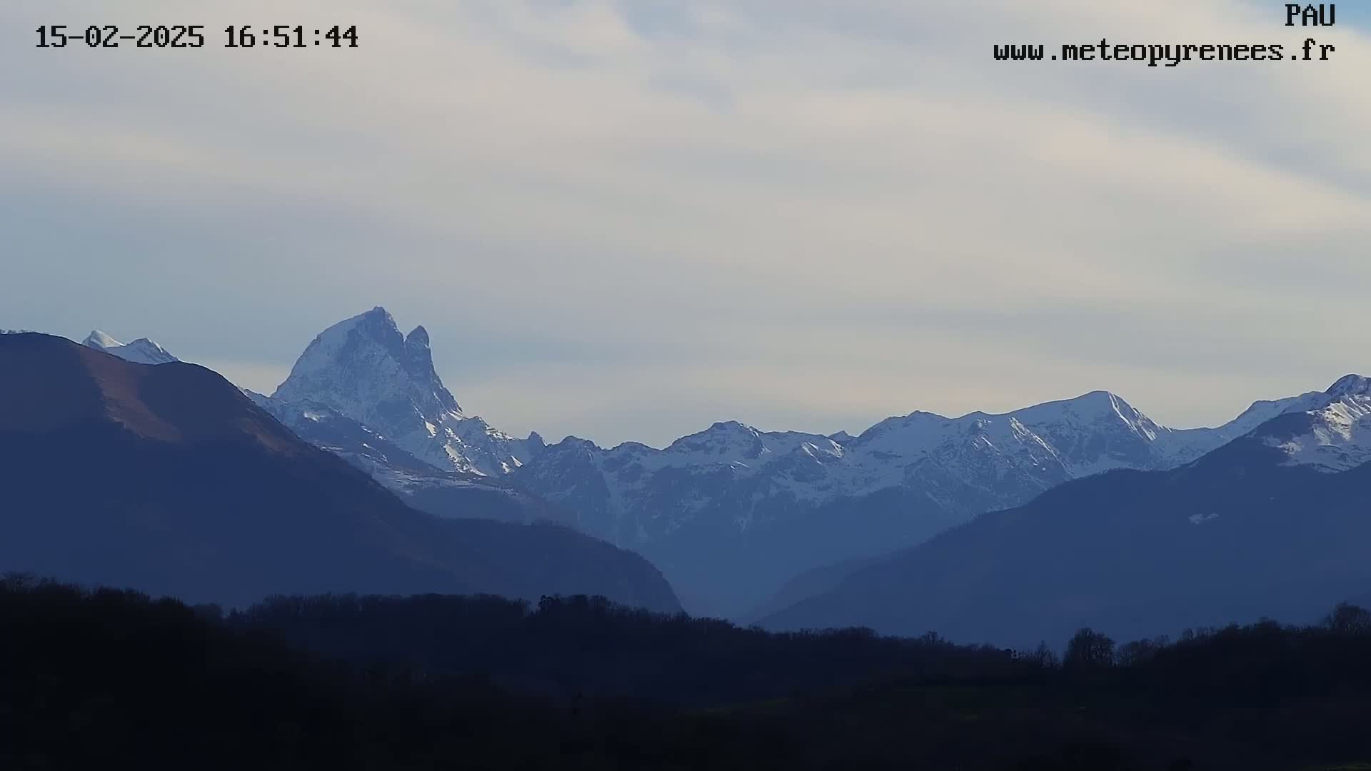 A snow-capped mountain range is visible in the distance under a partly cloudy sky, with dark silhouettes of trees and hills in the foreground.