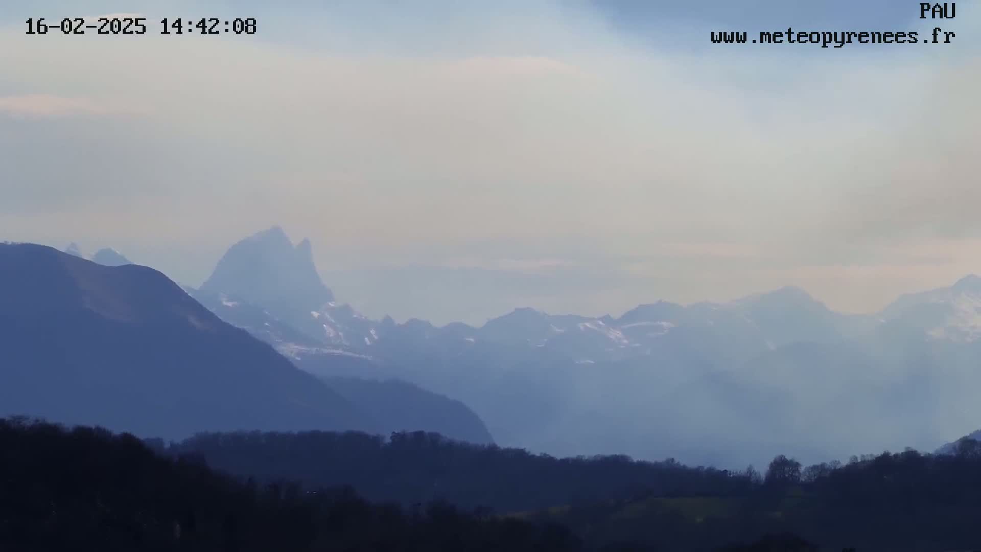 A hazy view of a mountain range, with snow visible on the peaks, stretches into the distance over a foreground of dark hills and trees.
