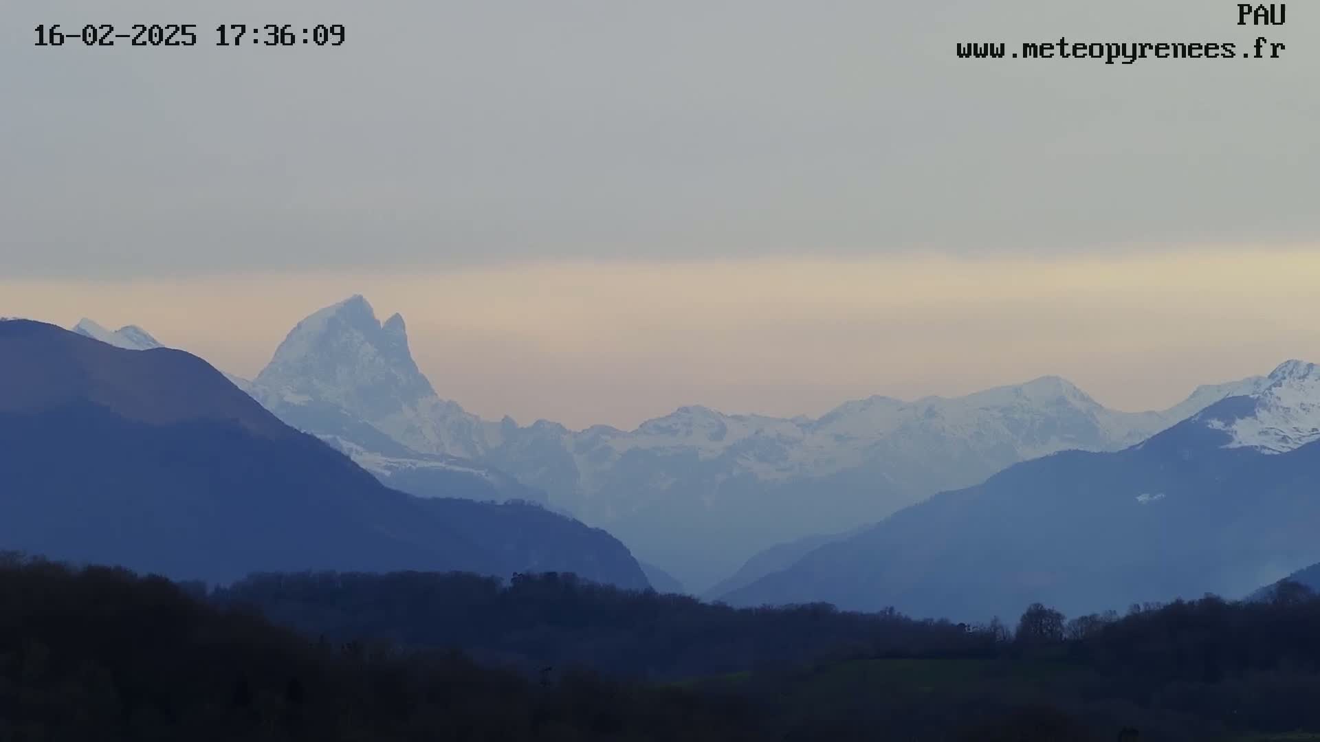 Snow-capped mountains rise in the distance under a pale, overcast sky, with a layer of dark hills and trees in the foreground.