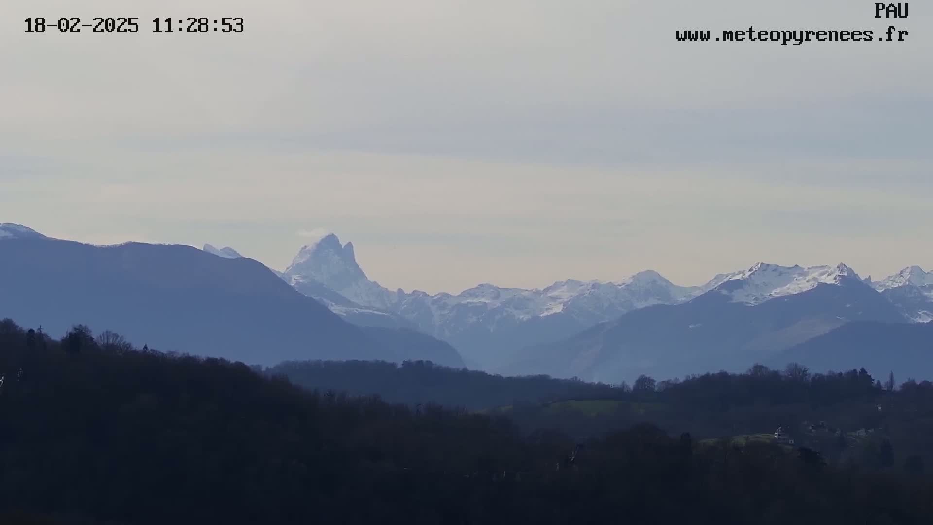 A snow-capped mountain range is visible in the distance, behind a foreground of dark, low-lying hills and trees under a mostly clear sky.