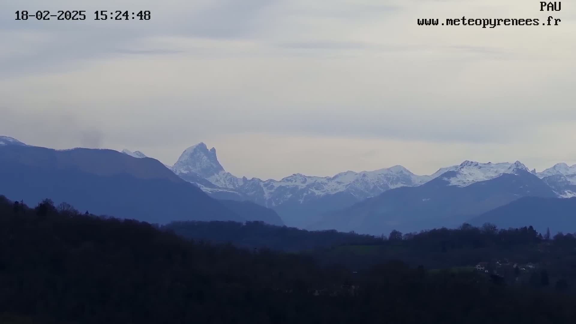 A long shot shows a range of snow-capped mountains in the distance, with a layer of dark trees and hills in the foreground under a mostly cloudy sky.