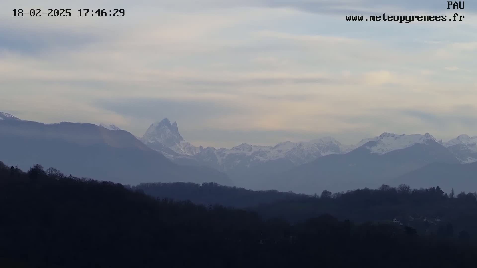 A hazy, overcast sky hangs over a distant, snow-capped mountain range behind a layer of dark, silhouetted hills and trees.