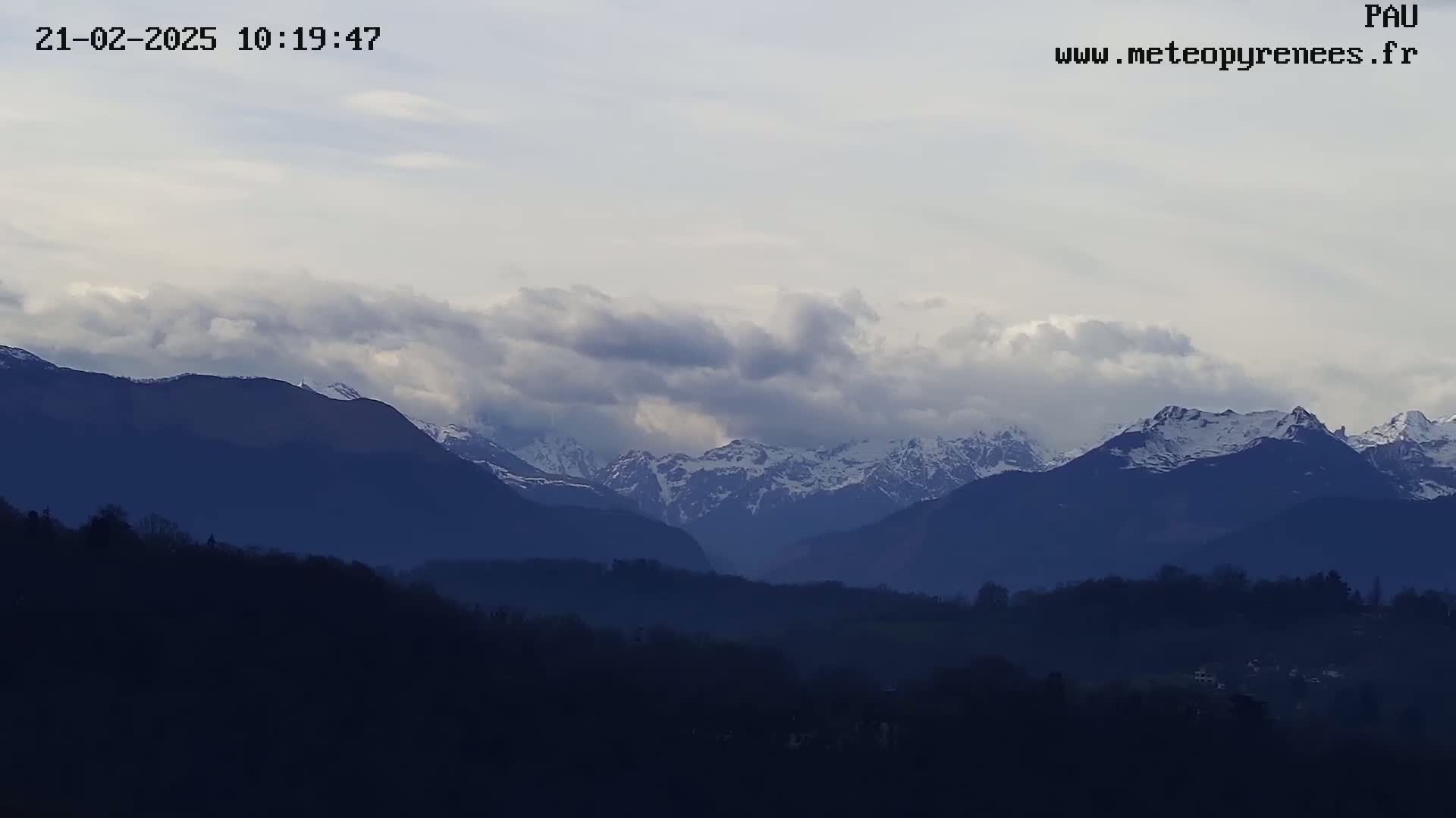 Snow-capped mountains are visible in the distance under a cloudy sky, with a foreground of dark, silhouetted hills and trees.