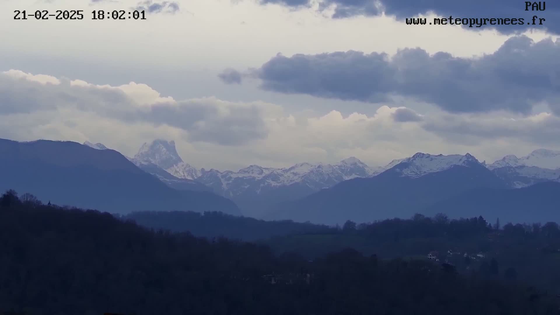 A partly cloudy sky overlooks a vast landscape of rolling hills and snow-capped mountains in the distance.