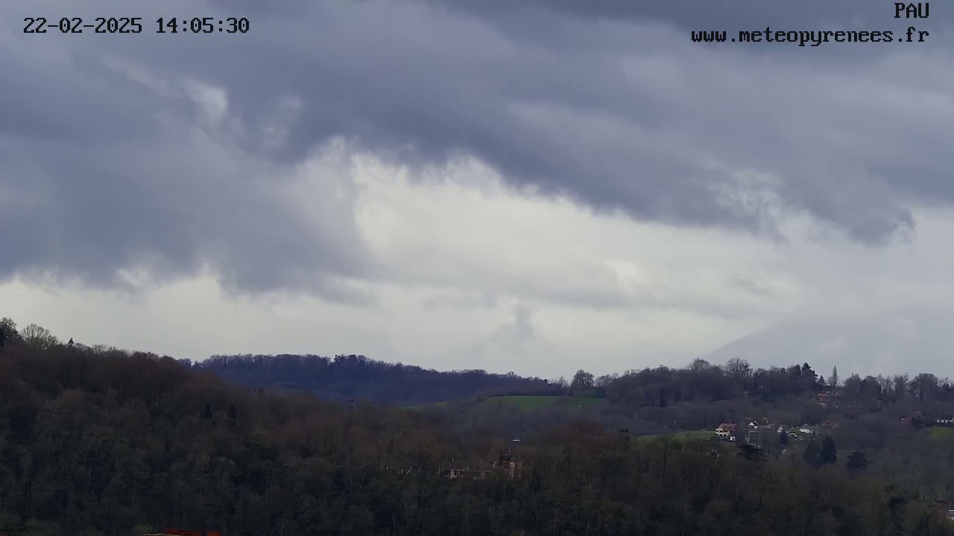 Overcast skies loom over a hilly landscape covered in dark, leafless trees and a few scattered buildings.