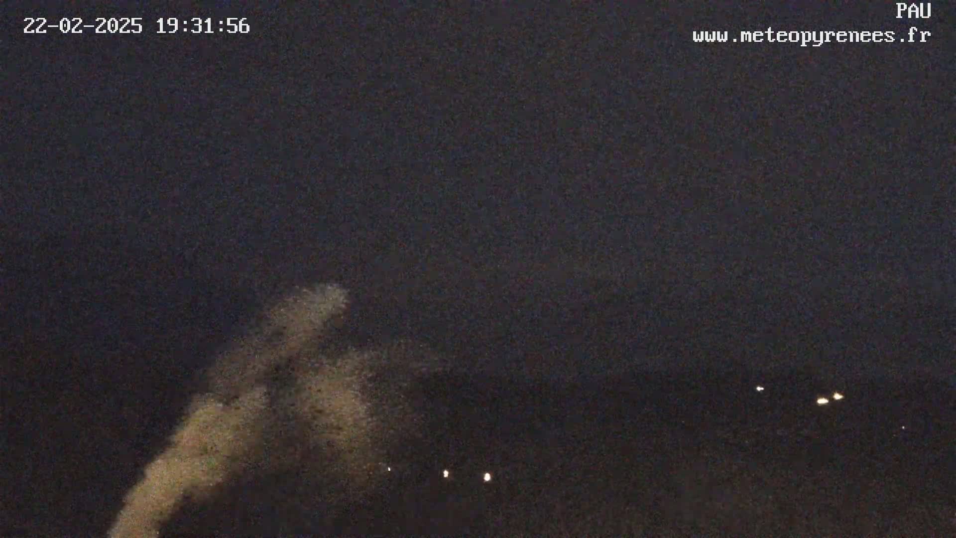 A nighttime outdoor scene shows a faint, light-brown plume rising against a dark sky with a few distant lights visible on the horizon.
