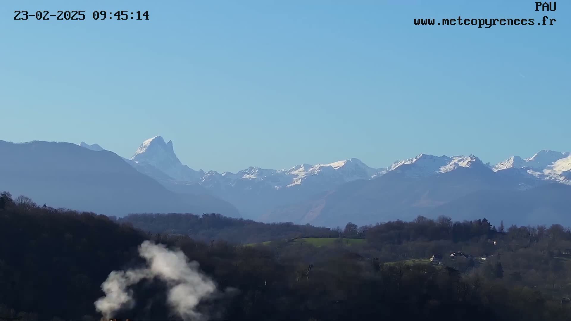A plume of smoke rises from a valley below a range of snow-capped mountains under a clear blue sky.