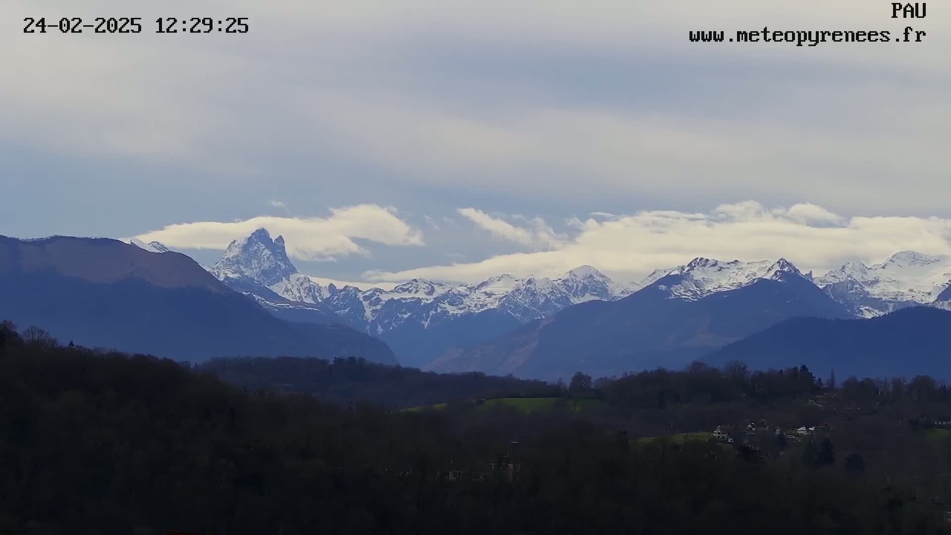 A long-range view shows a snow-capped mountain range under a partly cloudy sky, with a valley of dark trees and buildings in the foreground.