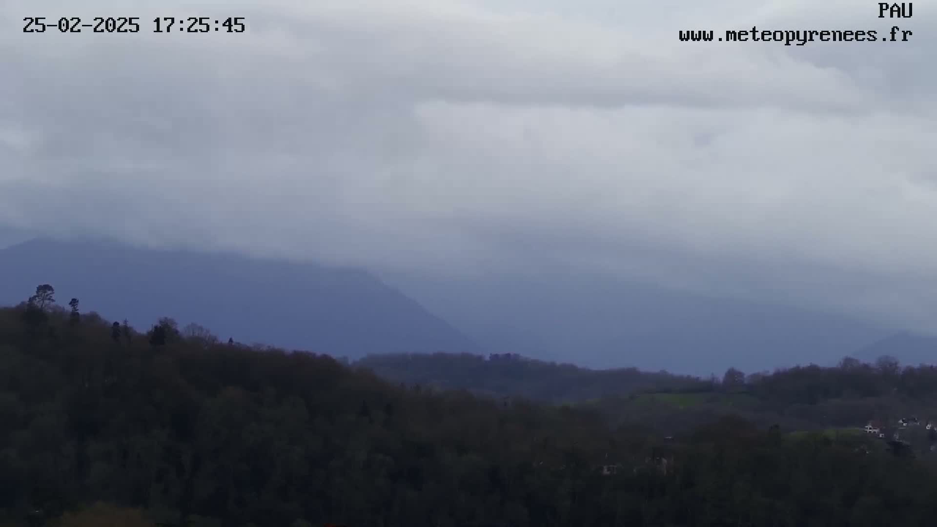 A view of dark, overcast skies over a hilly landscape covered in dark green trees and distant, hazy mountains.