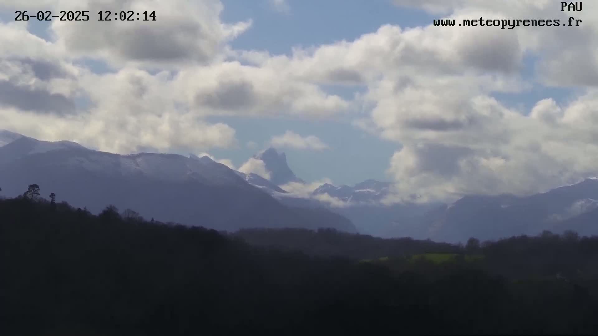 A partly cloudy sky overlooks a snow-capped mountain range in the distance, behind a dark foreground of hills and trees.