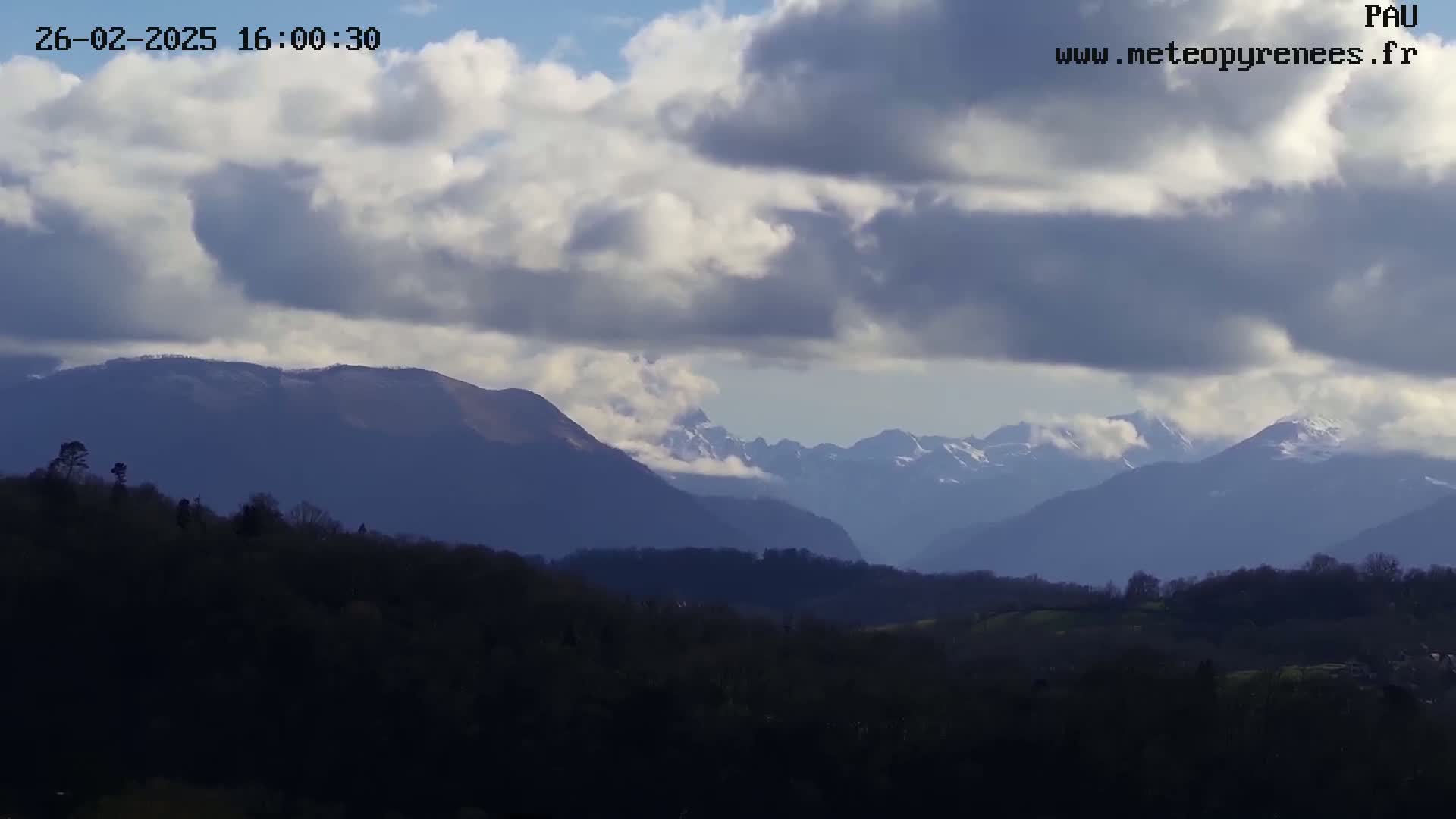 A partly cloudy day reveals a view of snow-capped mountains in the distance, with dark hills and trees in the foreground.