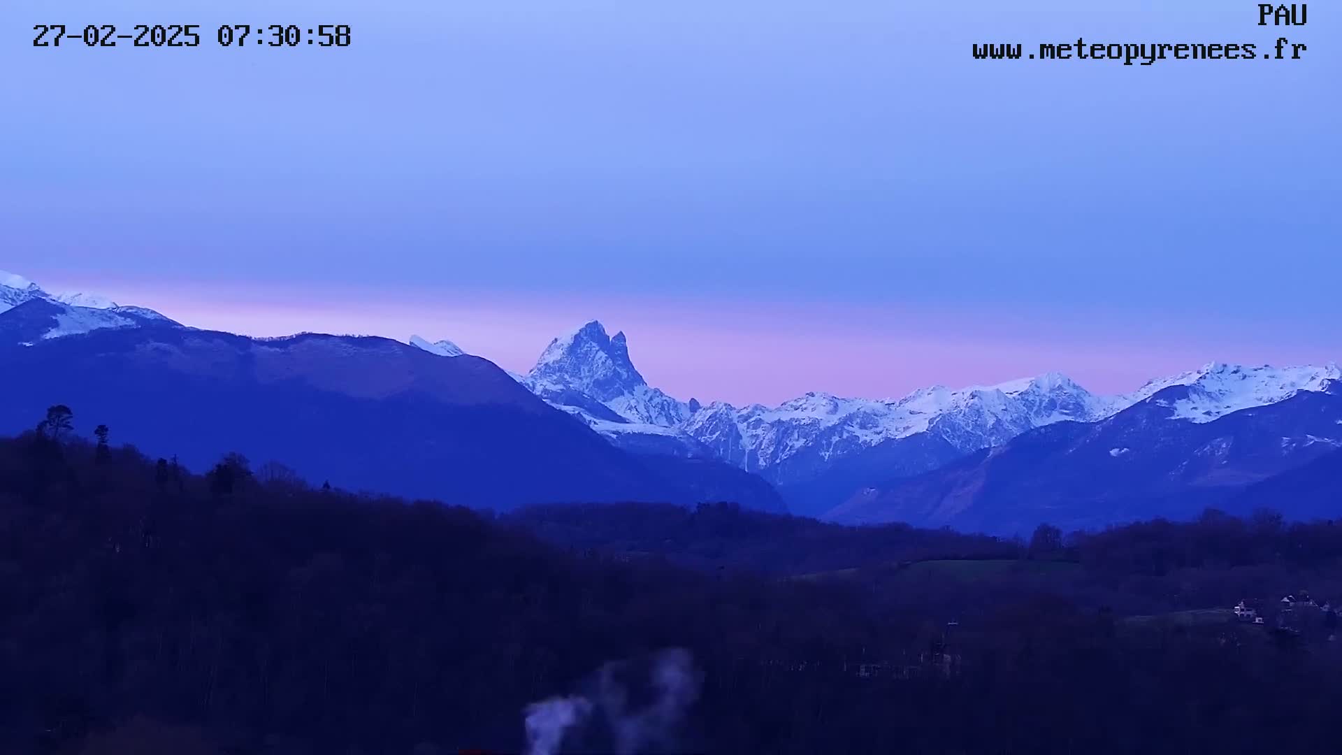 A snow-capped mountain range is visible at dawn under a clear, purplish sky, with a dark foreground of trees and low hills.
