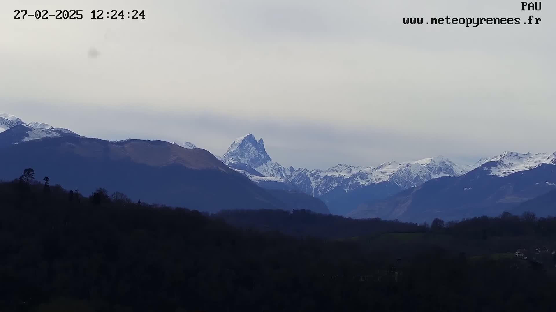 A distant view of snow-capped mountains under a mostly cloudy sky, with dark, silhouetted hills and trees in the foreground.
