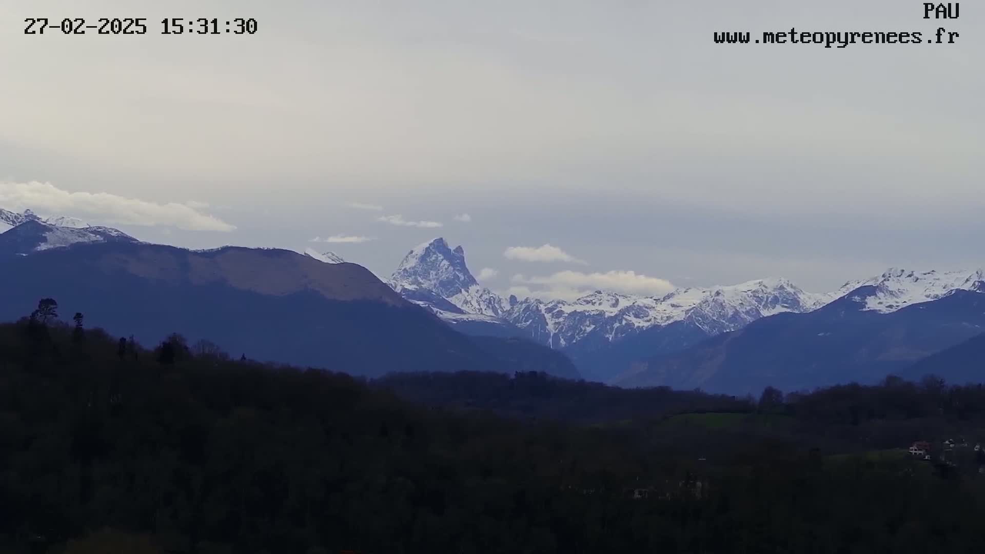 A snow-capped mountain range is visible in the distance, under a partly cloudy sky, behind a foreground of dark, silhouetted hills and trees.