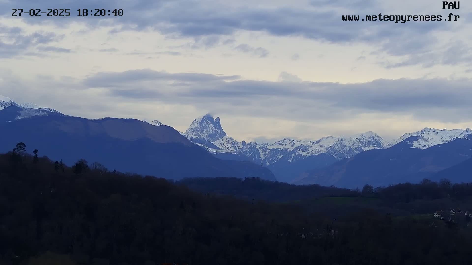 A view of snow-capped mountains in the distance, under a cloudy sky, with a foreground of dark, silhouetted trees and hills.