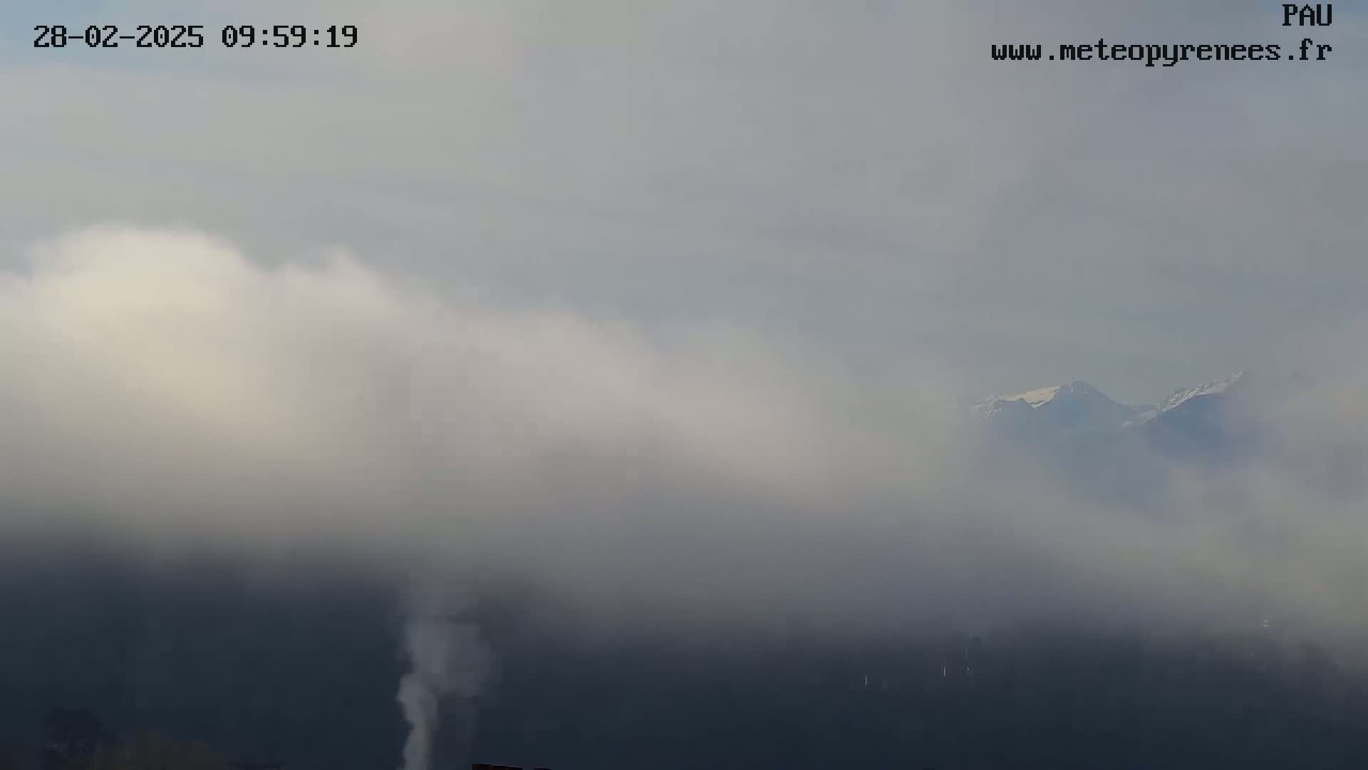 A hazy, foggy scene shows snow-capped mountains partially obscured by low-lying clouds in the distance.