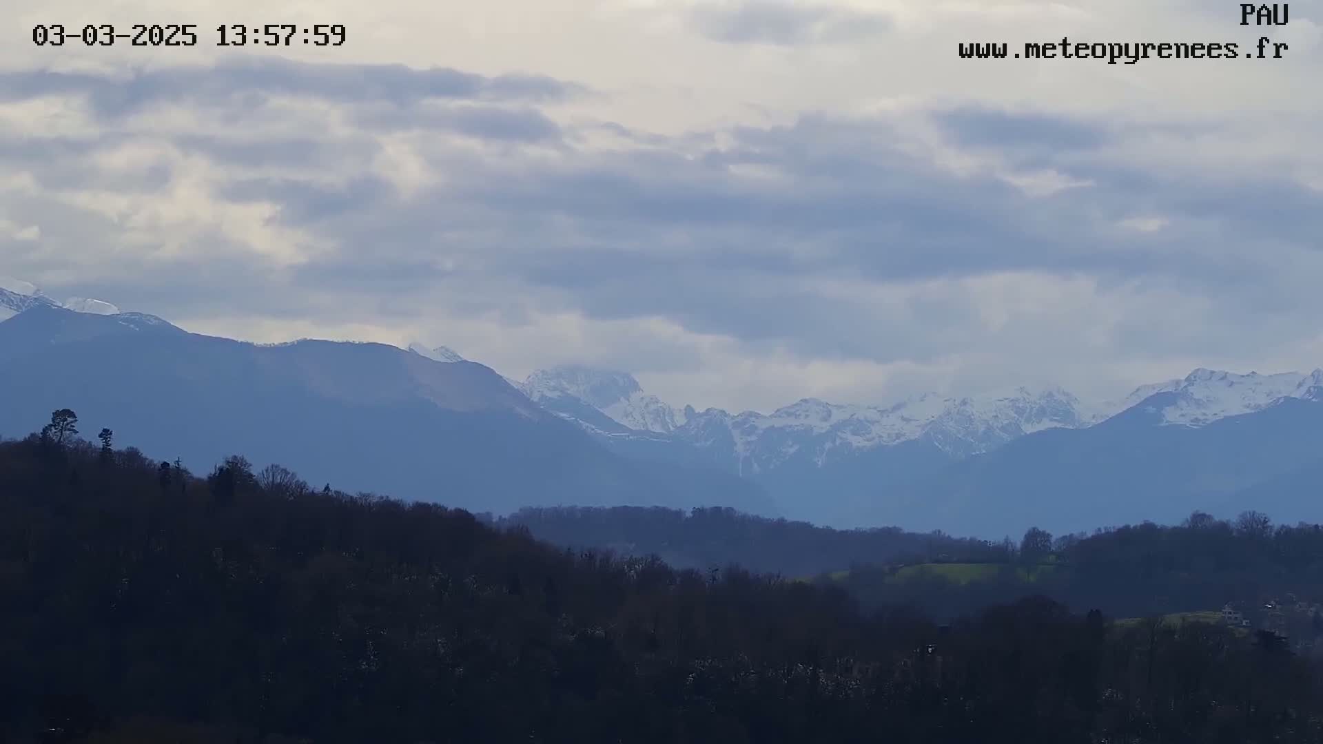 A mostly cloudy sky overlooks a range of snow-capped mountains in the distance, with a foreground of dark, low-lying hills and trees.