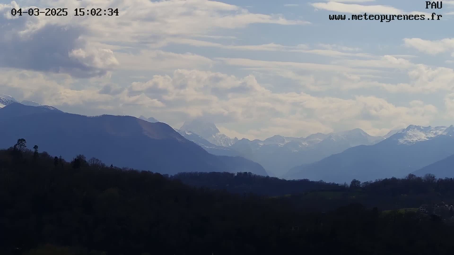 A partly cloudy sky overlooks a range of dark blue mountains, some with snow-capped peaks, behind a foreground of dark, silhouetted trees and hills.