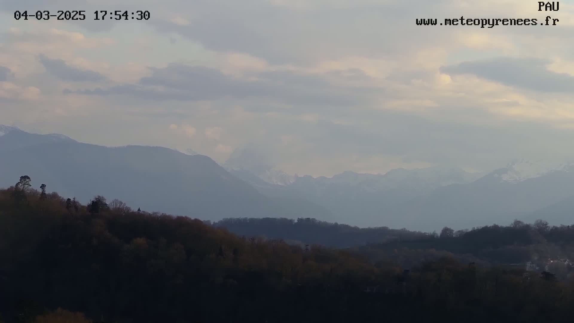 A partly cloudy sky overlooks a line of distant, hazy mountains and a foreground of dark, low-lying forest.