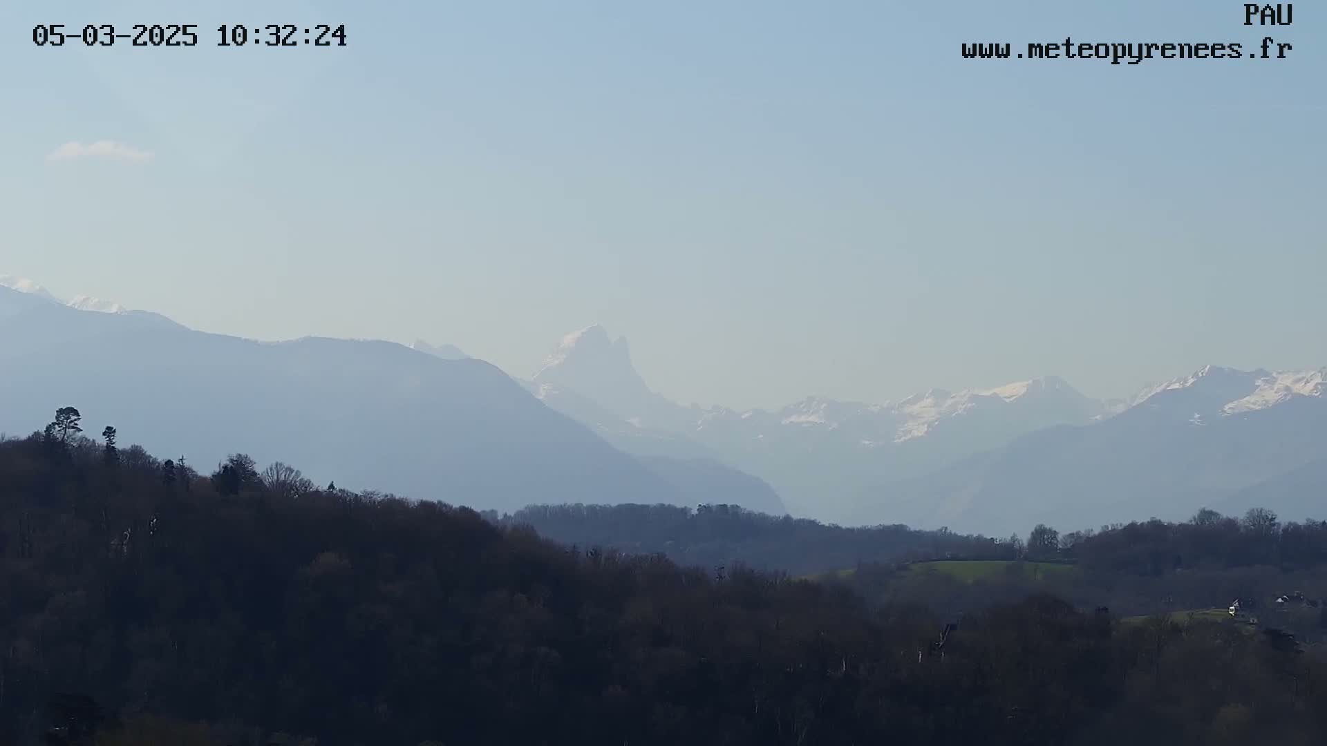 A hazy view of snow-capped mountains in the distance, over a landscape of dark green hills and trees under a clear, light blue sky.
