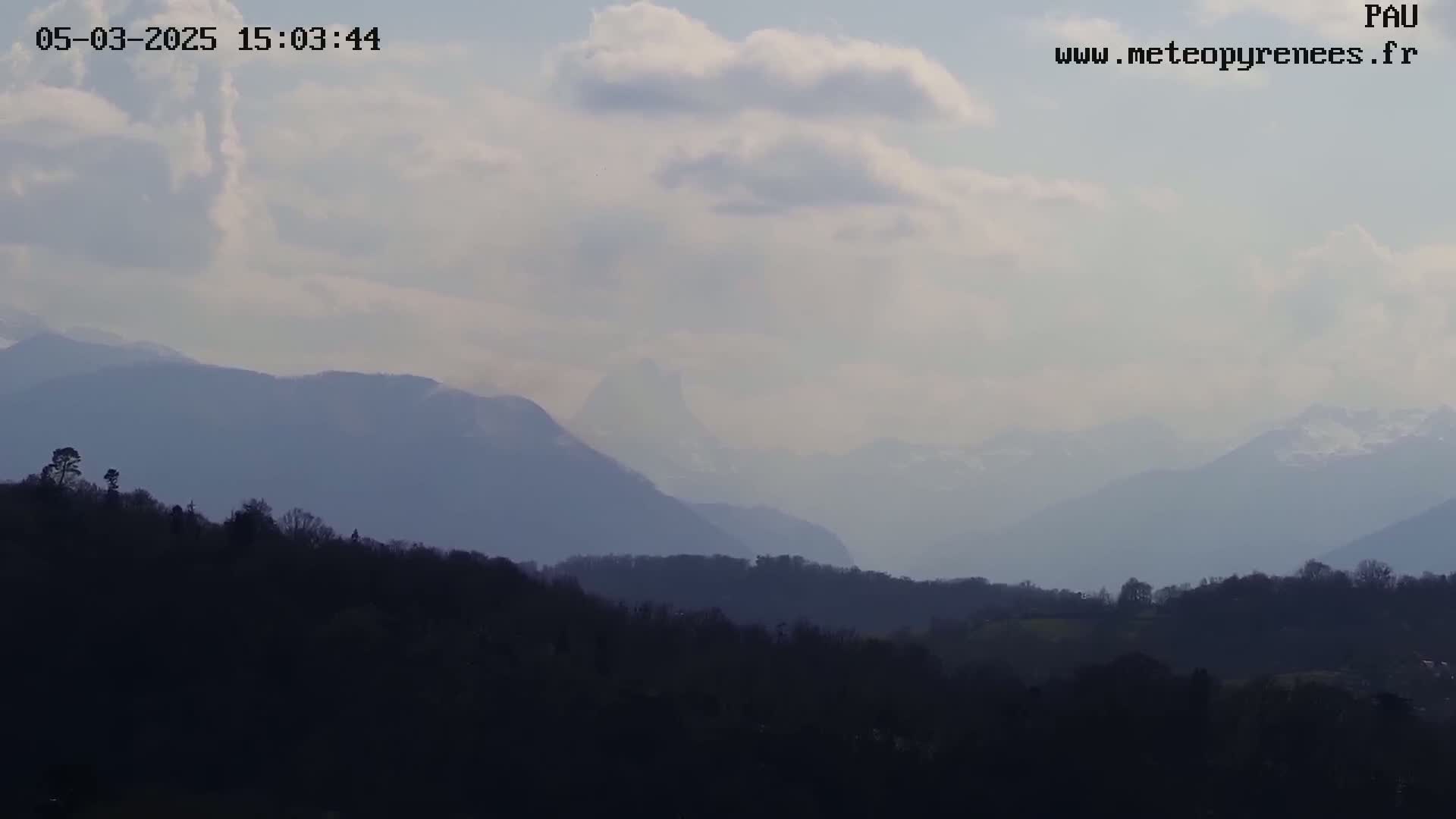 A hazy, partly cloudy day reveals a distant mountain range with snow-capped peaks behind a foreground of dark, tree-covered hills.